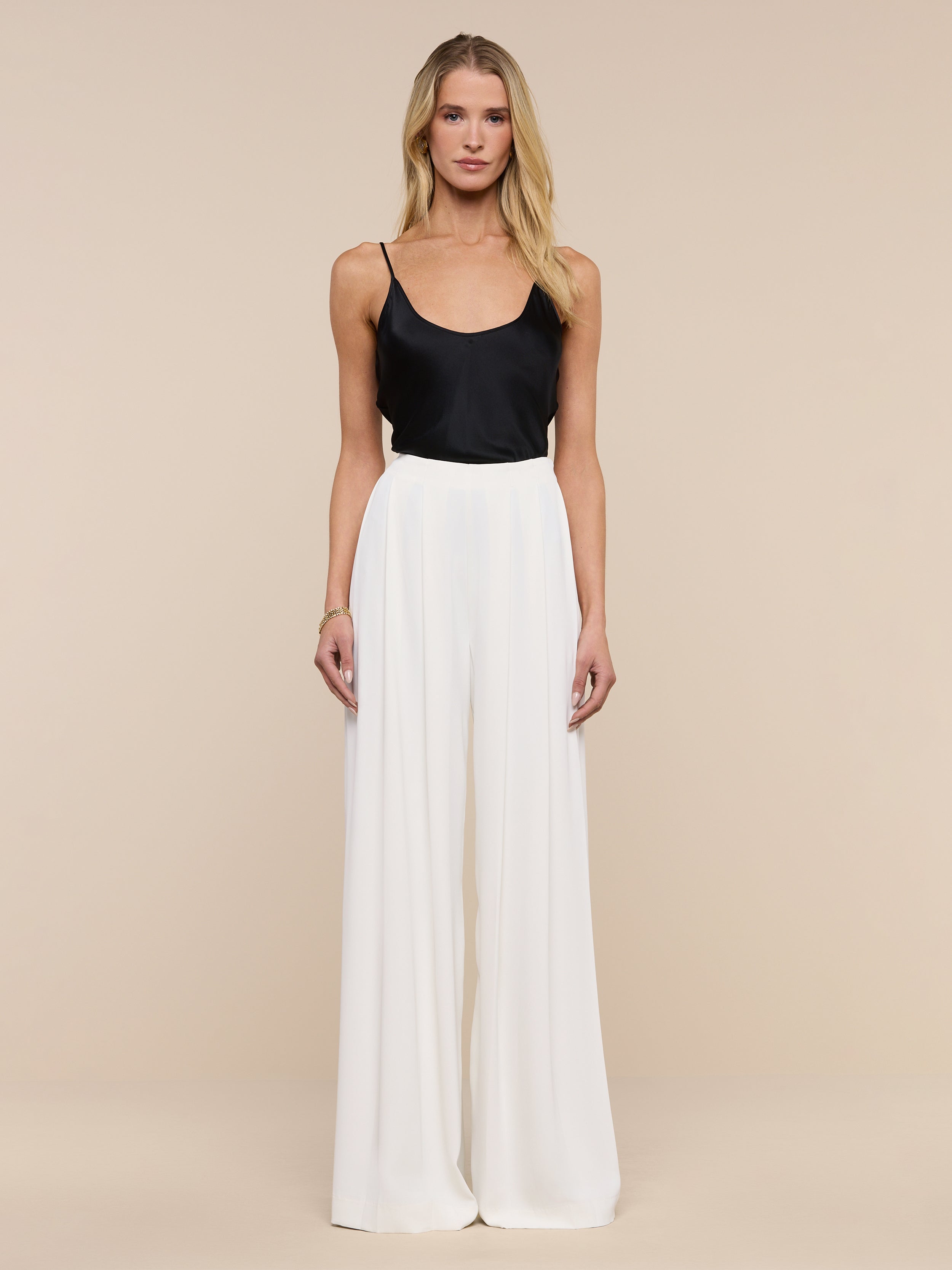 A woman stands facing forward against a plain white background, wearing a black sleeveless top paired with the Nova Pleated Wide-Leg Pant in ivory.