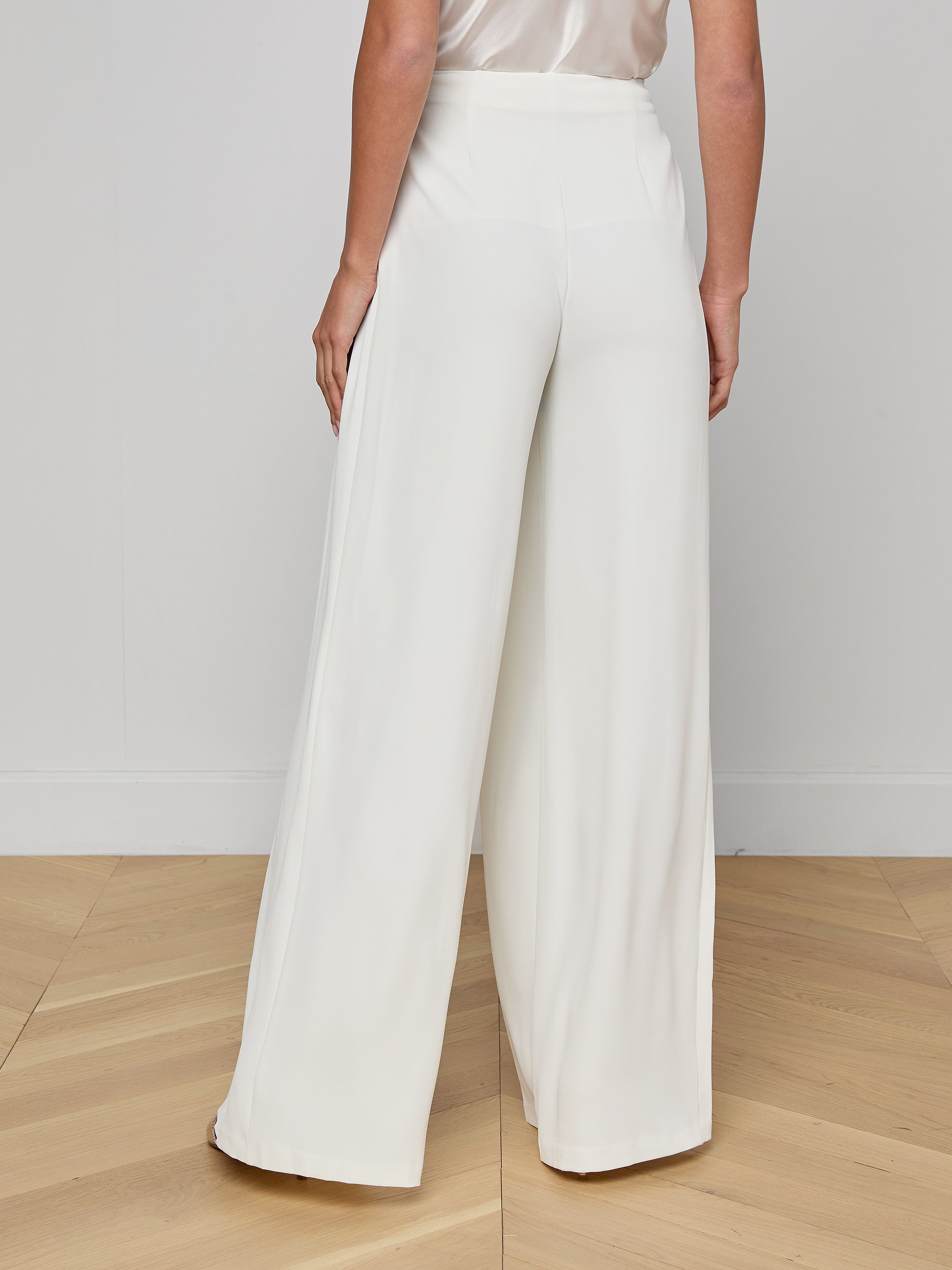 A person is shown from the shoulders down, standing against a plain white background, wearing the Nova Pleated Wide-Leg Pant in ivory and a sleeveless black top.