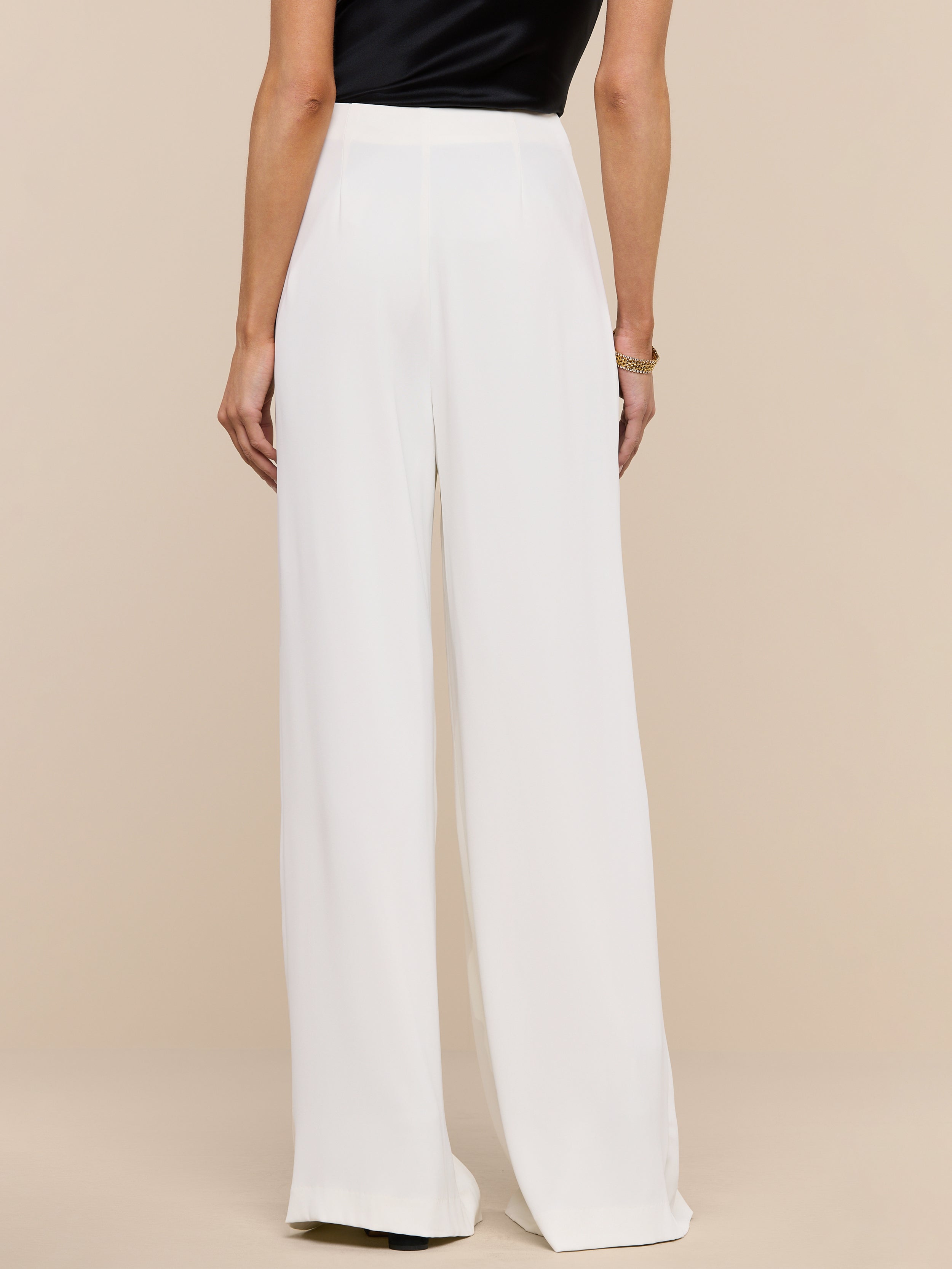 A person stands facing away, wearing the Nova Pleated Wide-Leg Pant in ivory paired with a sleeveless black top against a plain white background.