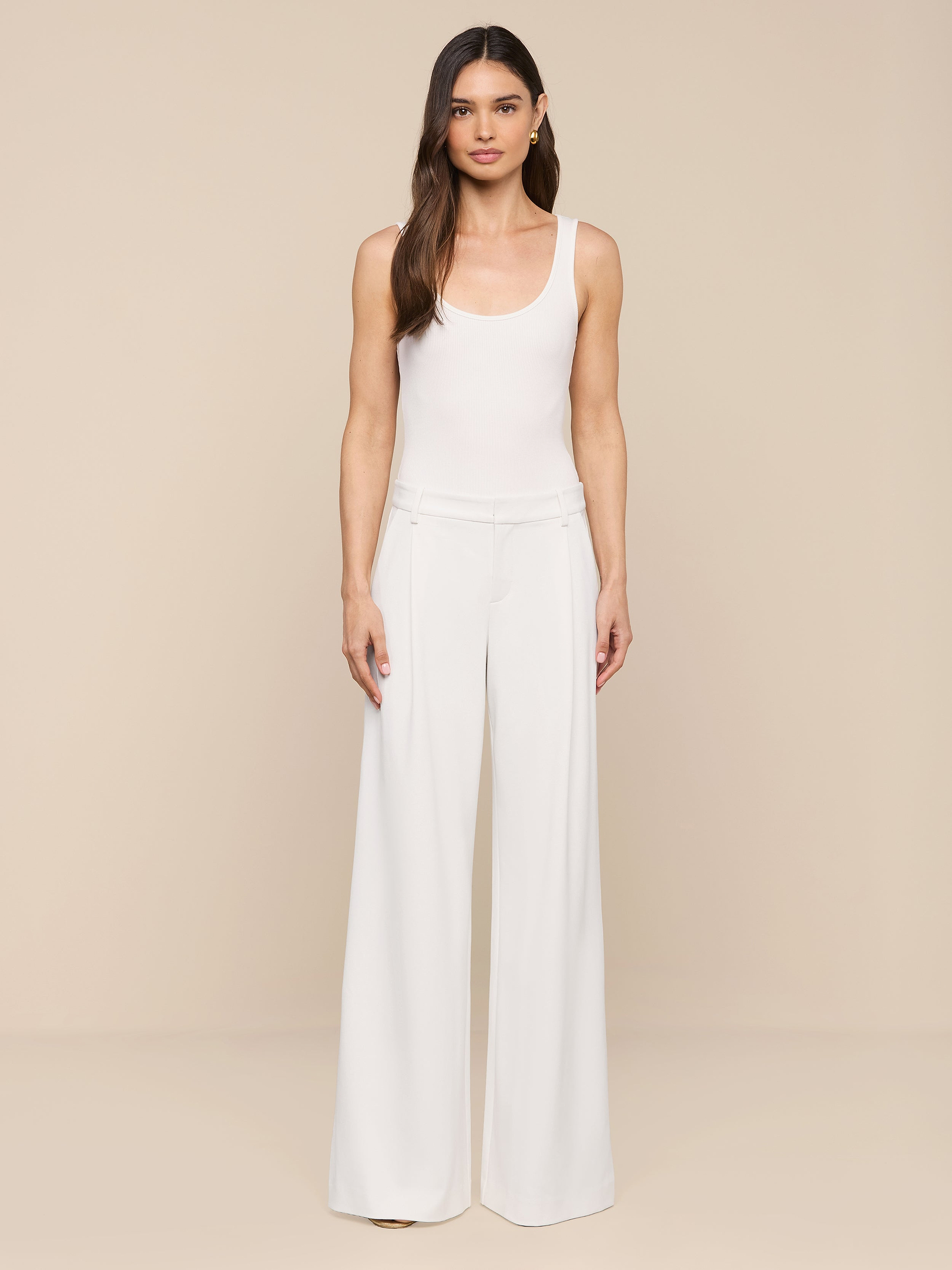 A woman with long brown hair and a neutral expression stands against a plain white background, wearing modern, wide-leg Allanah Trousers crafted from stretch suit fabric for a flattering fit, paired with a sleeveless white top.