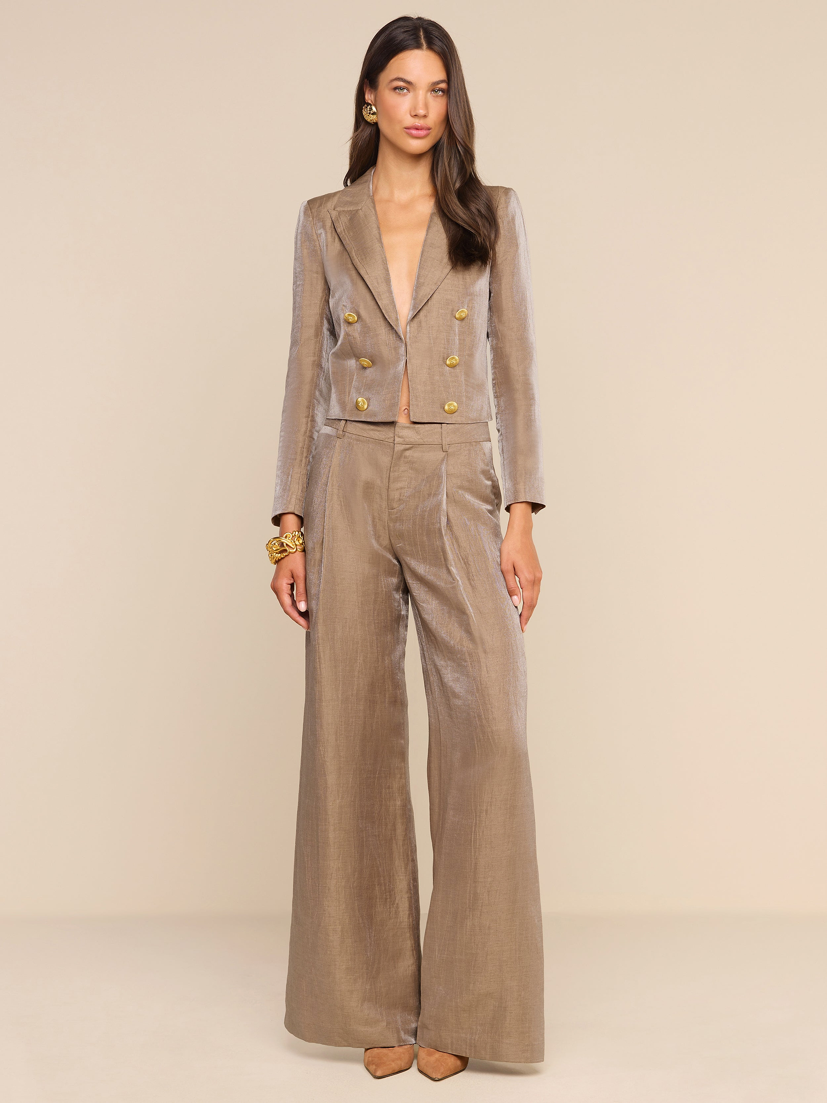 A woman stands against a plain white background wearing the Allanah Linen-Blend Trouser paired with a matching tan double-breasted blazer featuring gold buttons. She accessorizes with gold earrings, a gold bracelet, and pointed-toe heels, and wears her long hair down.