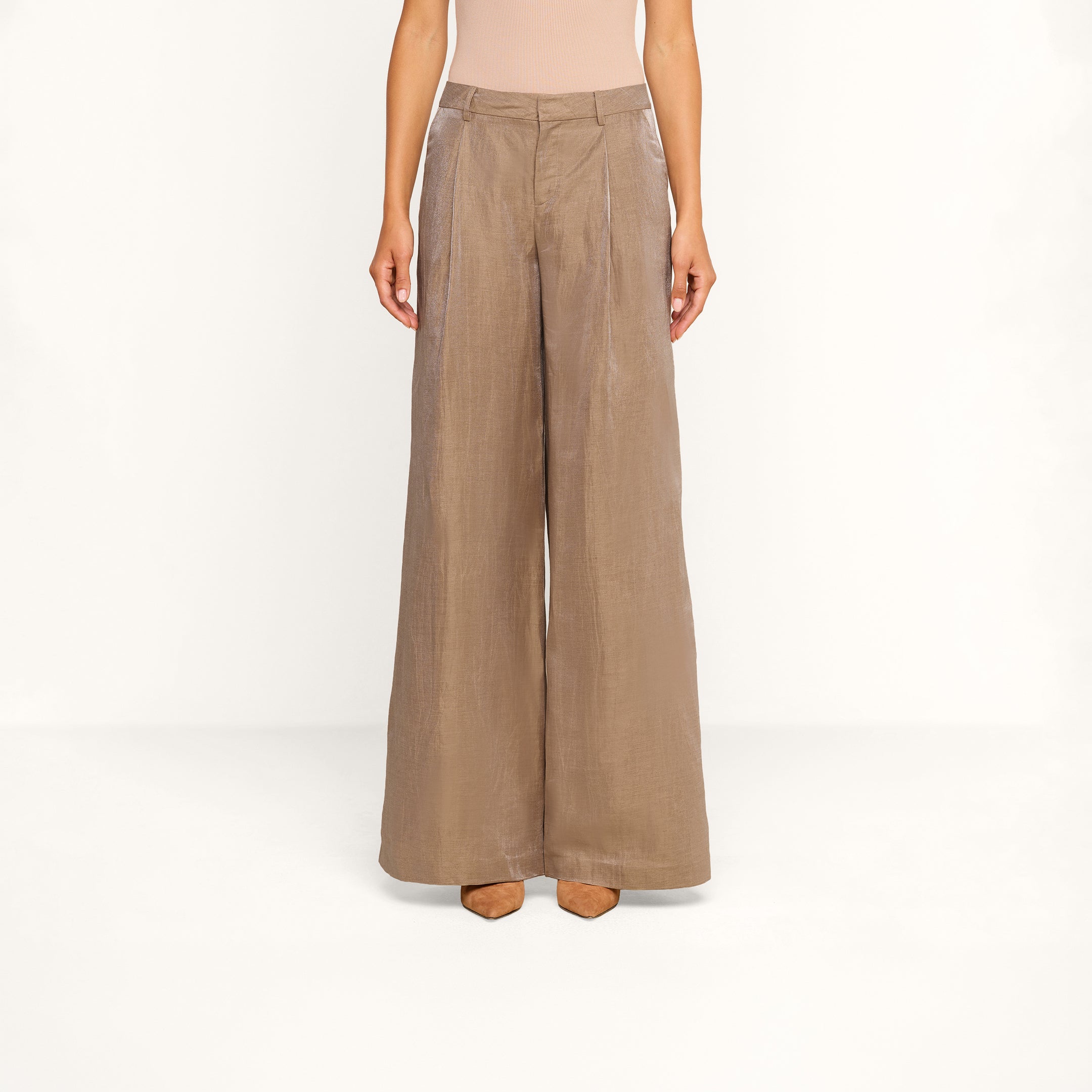 Una persona indossa i pantaloni Allanah in misto lino, a vita alta e a gamba larga, color marrone latte, abbinati a un top senza maniche beige e a scarpe con tacco marroni, in piedi davanti a uno sfondo beige.