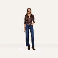 A woman wearing sunglasses, a dark brown long-sleeve shirt, the Scottie Wide-Leg Jean crafted from super stretch denim, a belt, and brown boots stands against a plain light background.