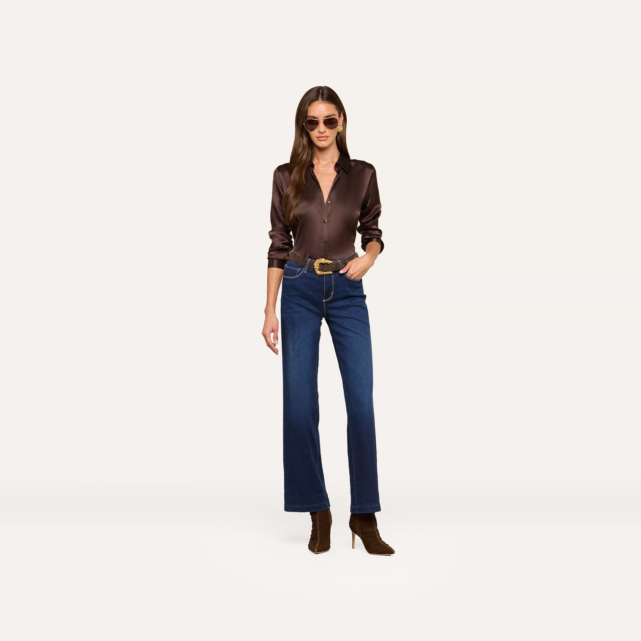 A woman wearing sunglasses, a dark brown long-sleeve shirt, the Scottie Wide-Leg Jean crafted from super stretch denim, a belt, and brown boots stands against a plain light background.