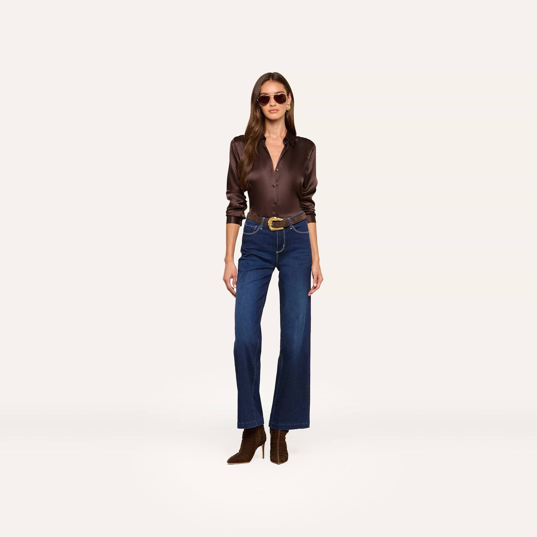Woman wearing sunglasses, a dark brown satin button-up shirt, the Scottie Wide-Leg Jean in super stretch denim, a brown belt, and brown ankle boots stands against a plain white background.