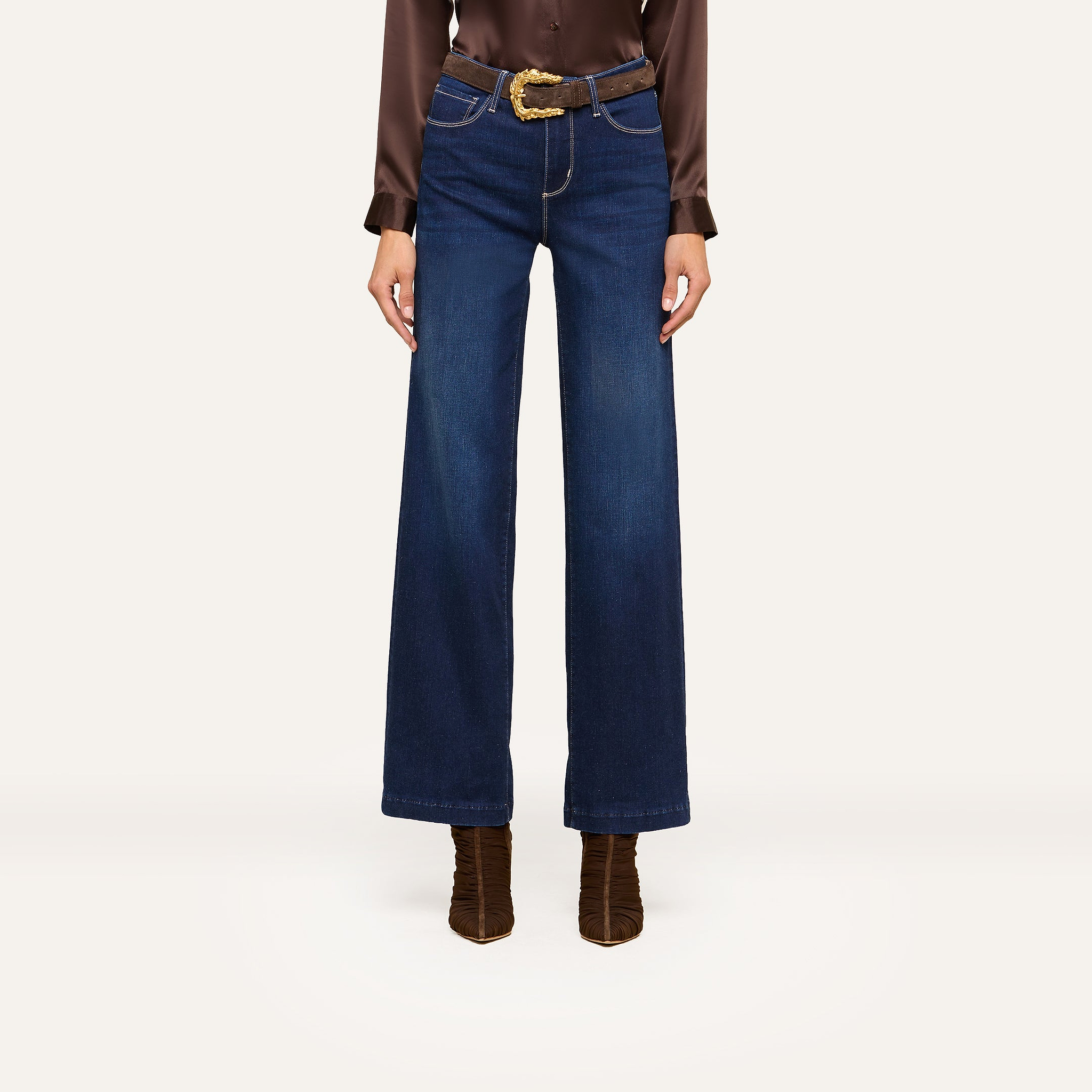 A person wearing high-waisted, wide-leg blue jeans and a brown long-sleeve blouse styled with a brown belt featuring a gold buckle and brown heeled boots stands confidently against a white background.