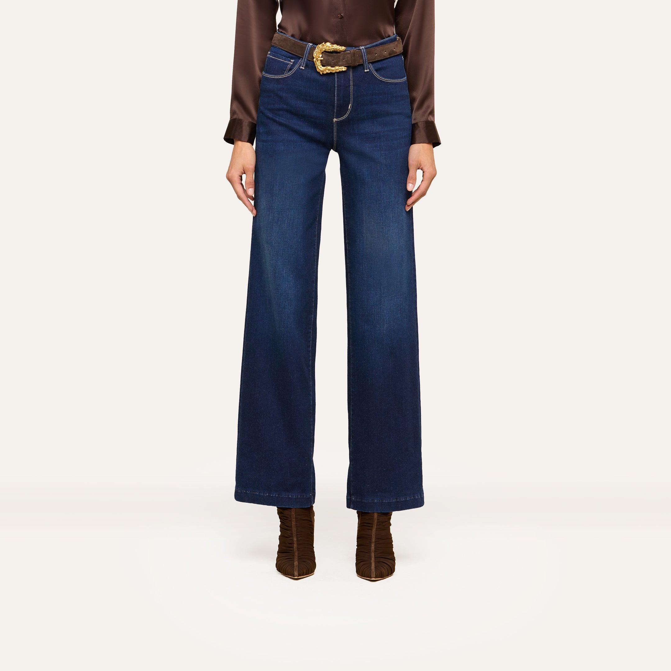 A person wearing high-waisted, wide-leg blue jeans and a brown long-sleeve blouse styled with a brown belt featuring a gold buckle and brown heeled boots stands confidently against a white background.