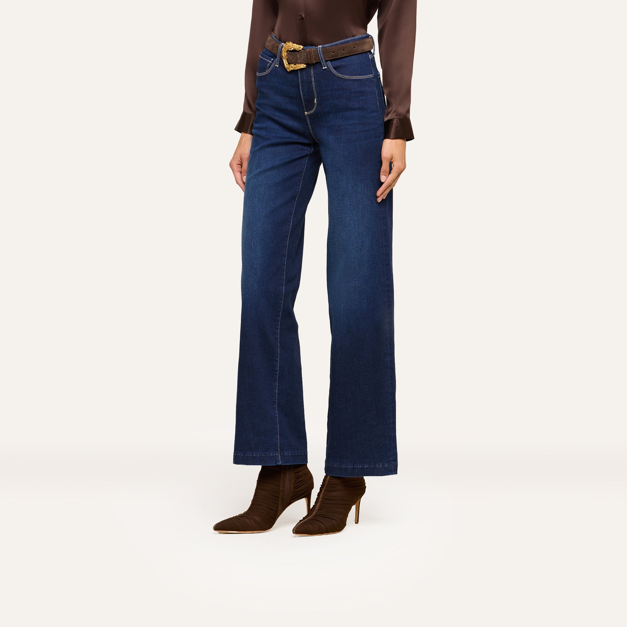 A person wearing a brown satin blouse, dark blue wide-leg jeans, a brown belt, and brown heeled boots stands against a plain white background.