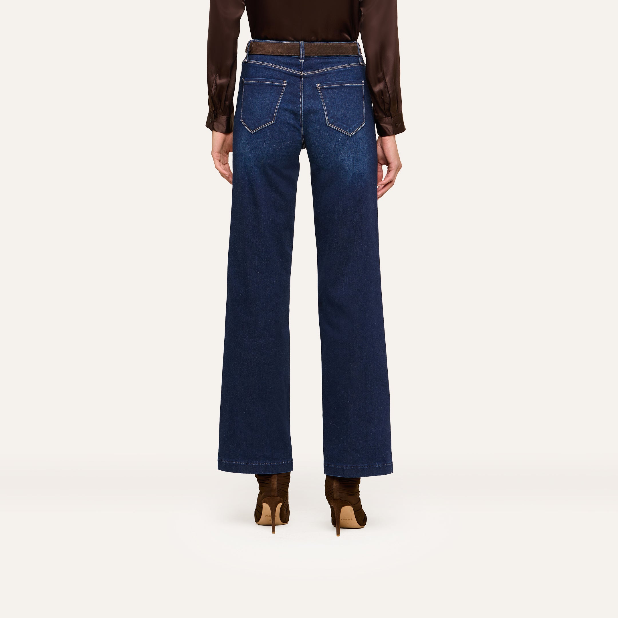 Person standing against a white background, shown from the back, wearing wide-leg dark blue jeans, a brown long-sleeve blouse, and stylish brown heeled boots.