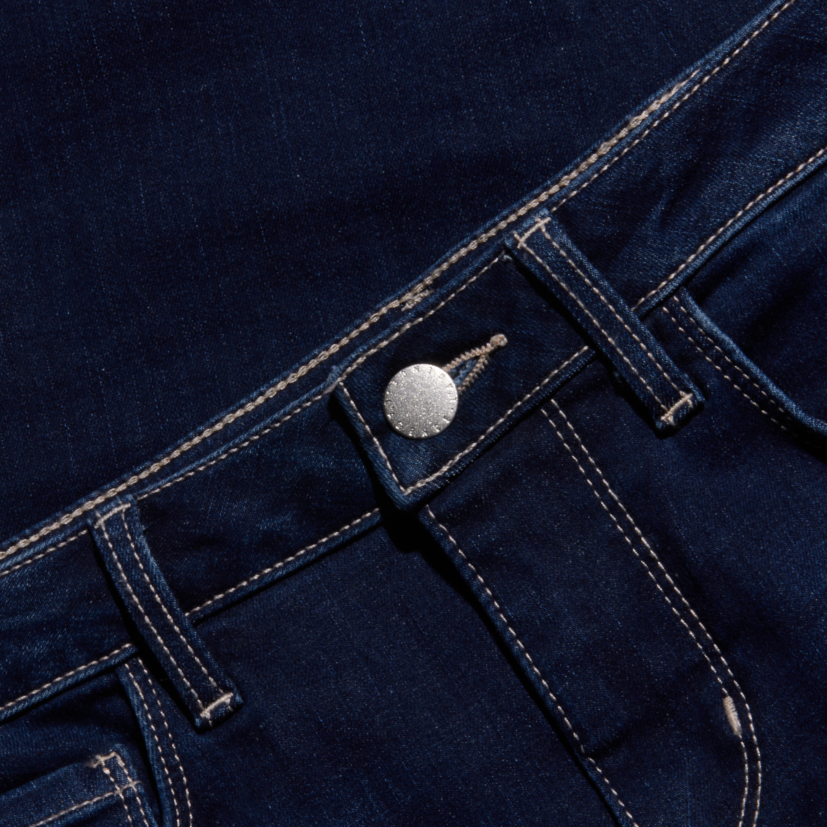 Close-up of the waistband and front pocket area of dark blue jeans with white stitching, silver button, and visible belt loops, set against a matching denim background.