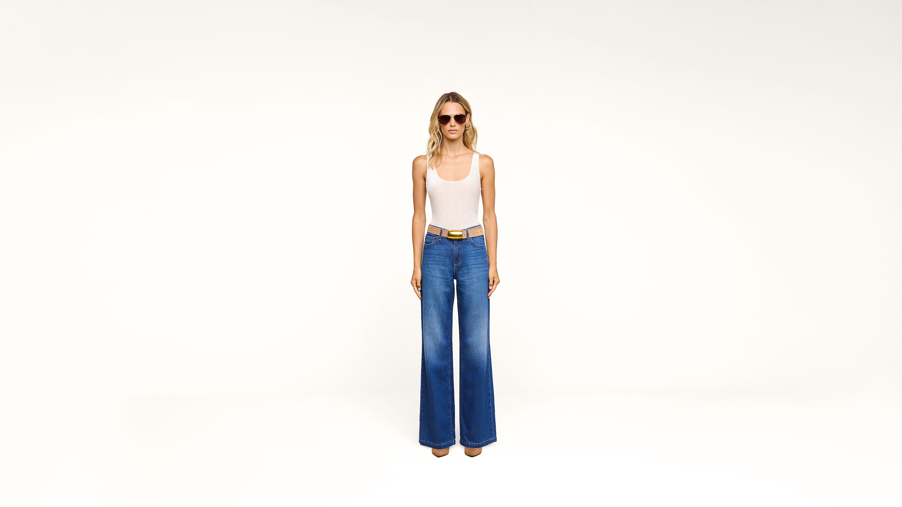 Woman stands against a plain white background, wearing sunglasses, a white tank top, blue high-waisted wide-leg jeans, and a tan belt.