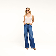 A woman wearing sunglasses, a white tank top, the Scottie Wide-Leg Jean in an indigo wash, and brown shoes stands against a plain white background with her hands in her pockets.