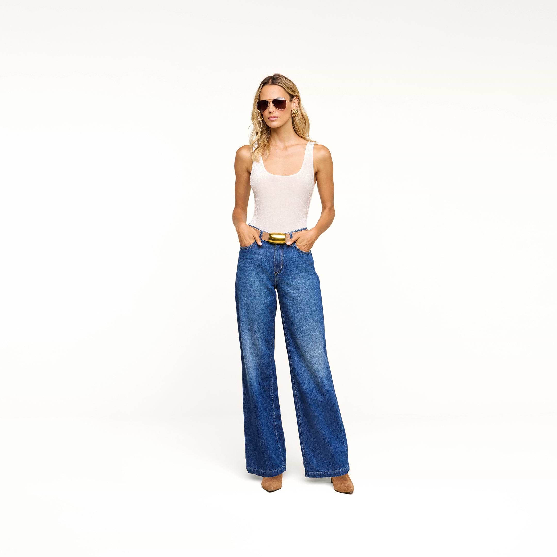 A woman wearing sunglasses, a white tank top, the Scottie Wide-Leg Jean in an indigo wash, and brown shoes stands against a plain white background with her hands in her pockets.