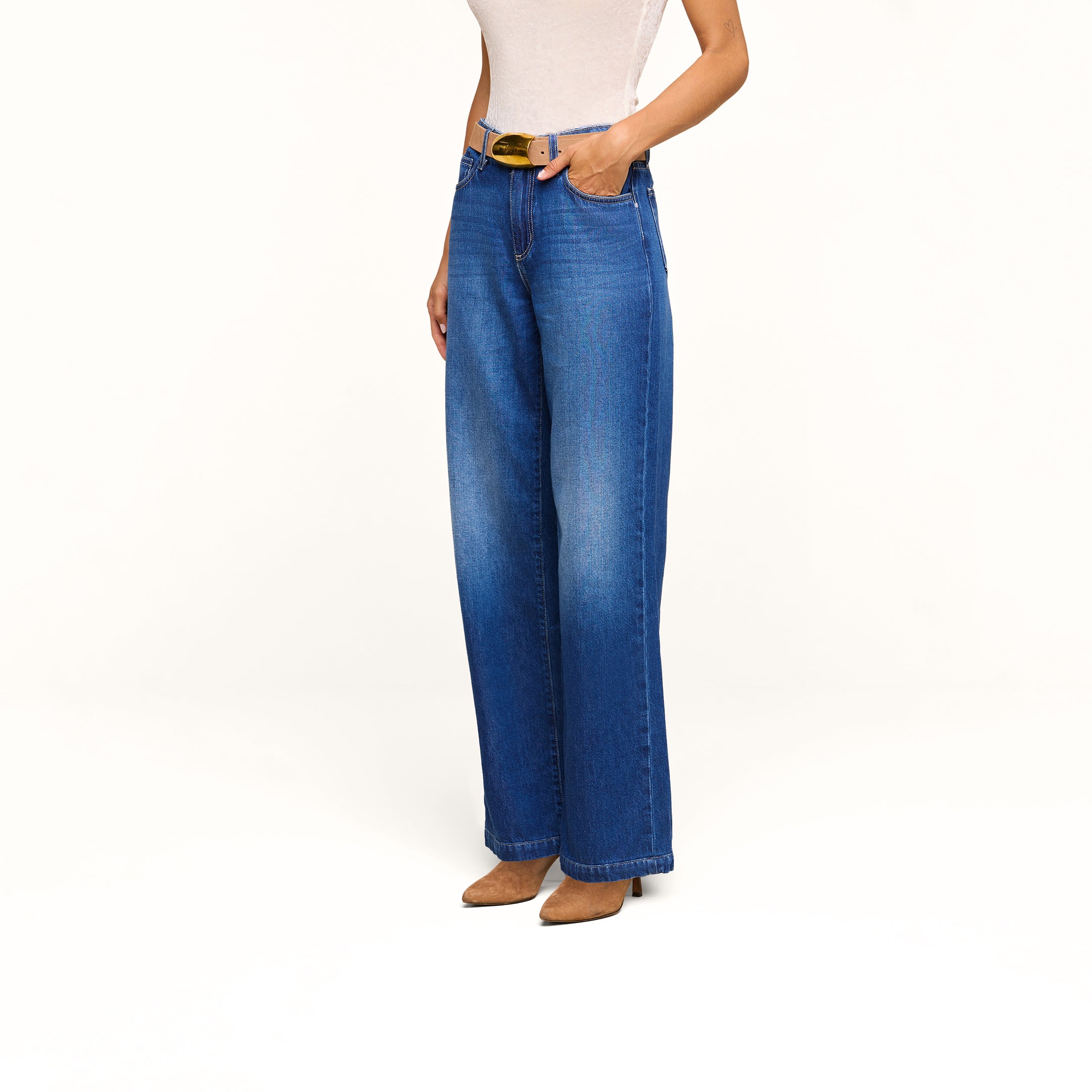 Woman wearing a sleeveless beige top, the Scottie Wide-Leg Jean in an indigo wash with a high-rise fit and black belt, paired with brown pointed-toe heeled shoes, standing against a plain white background.