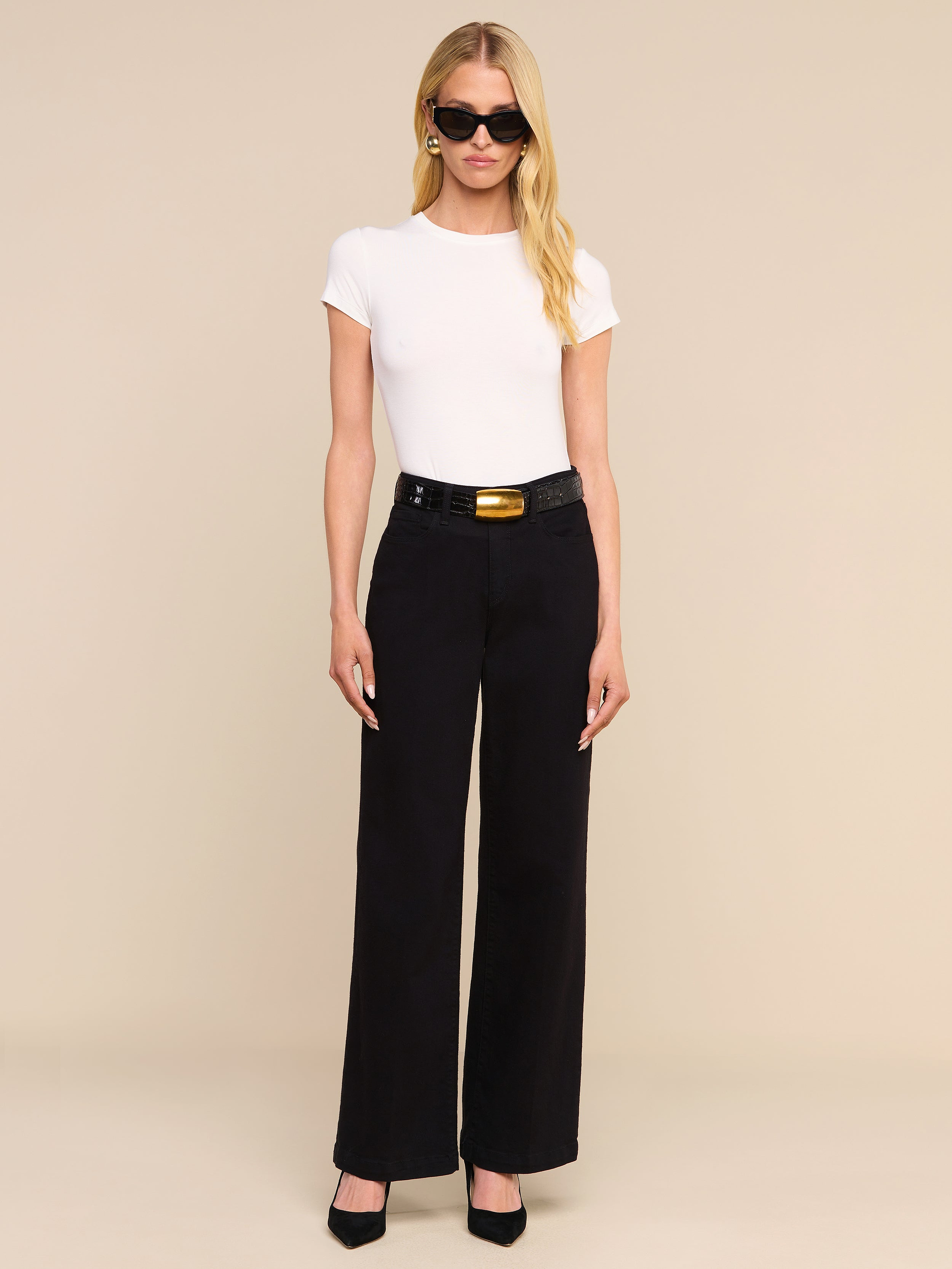 A woman with long blonde hair, wearing black sunglasses, a white short-sleeve top, and the Scottie Wide-Leg Jean paired with a black belt and black heels stands against a plain white background.