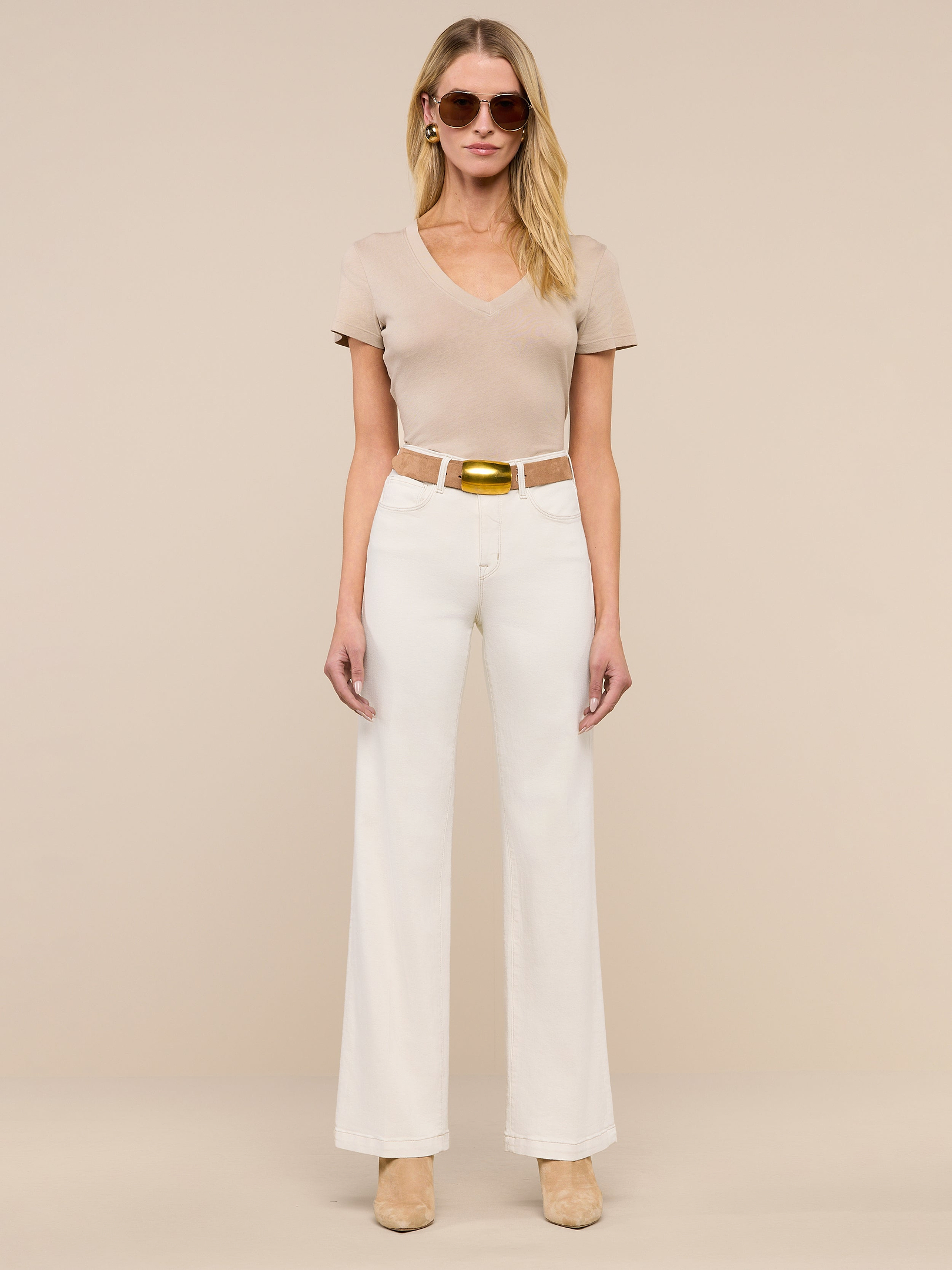 A woman wearing beige sunglasses, a short-sleeve beige top, the Scottie Wide-Leg Jean in cream with a tan belt, and beige heels stands against a plain light background.