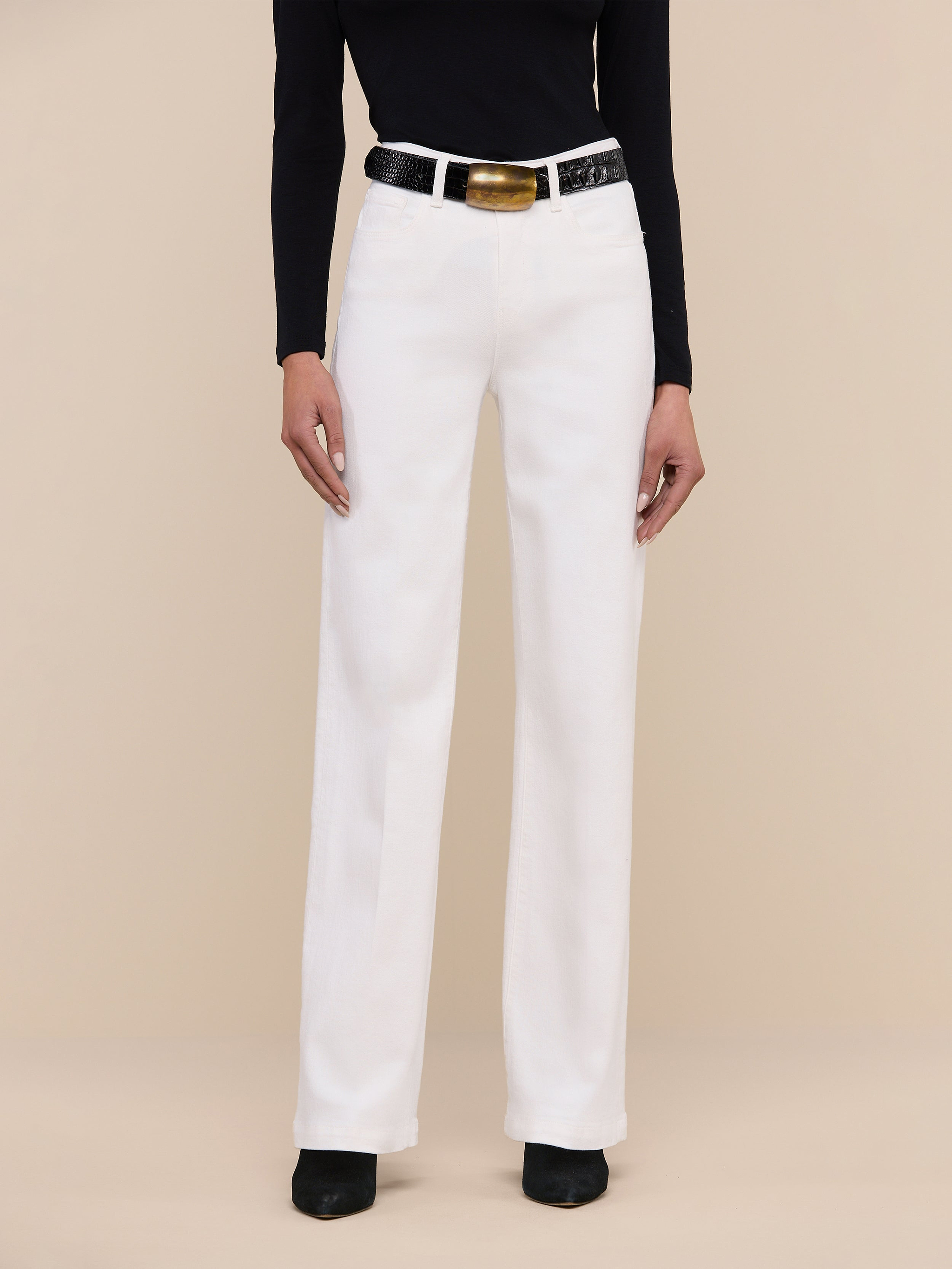 Wearing the Scottie Wide-Leg Jean in white, a person stands against a plain light background, styled with a black sleeveless top, a white belt with a gold buckle, and black pointed-toe shoes.