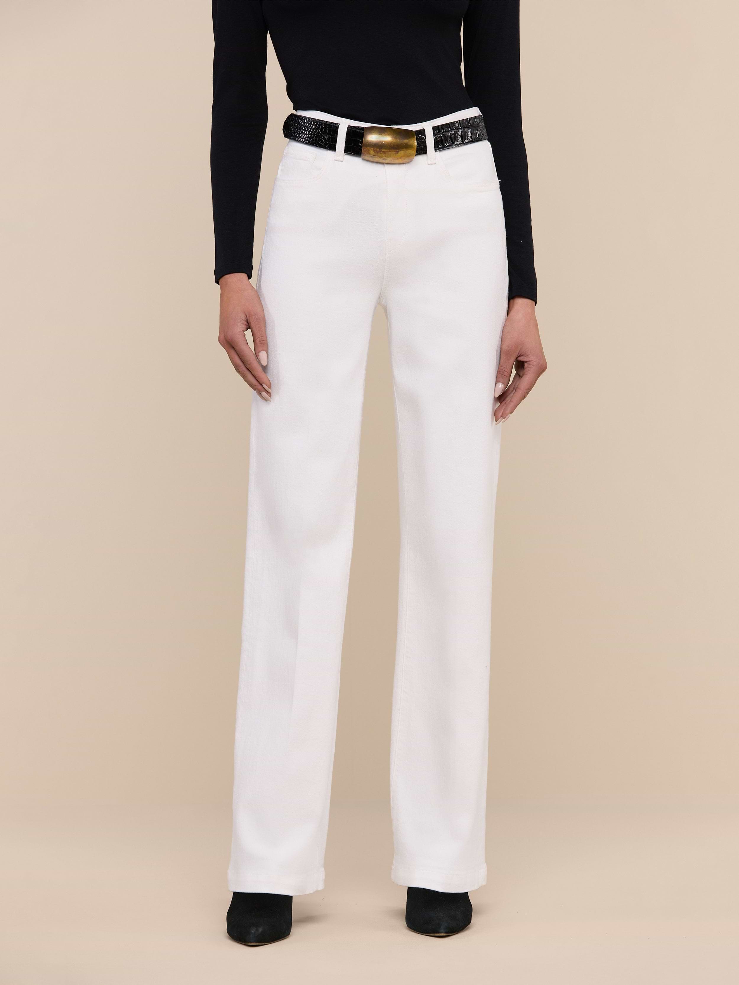 Wearing the Scottie Wide-Leg Jean in white, a person stands against a plain light background, styled with a black sleeveless top, a white belt with a gold buckle, and black pointed-toe shoes.