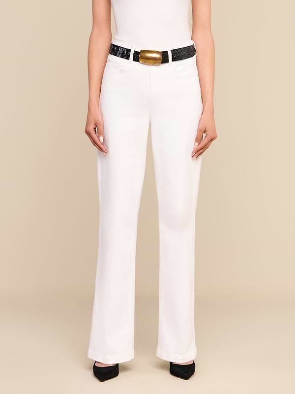 Una persona che indossa i jeans Scottie Wide-Leg di colore bianco, abbinati a un top bianco senza maniche, una cintura nera con una grande fibbia dorata e scarpe nere a punta, è in piedi su uno sfondo beige.