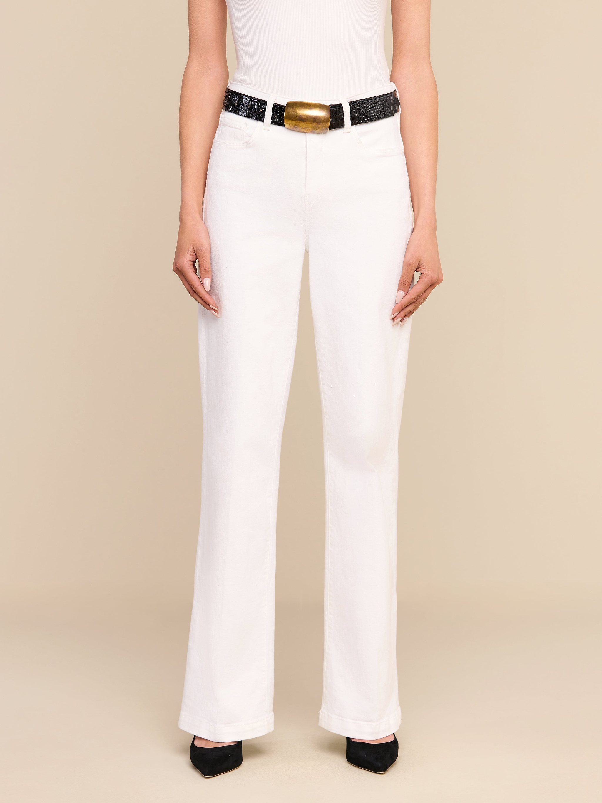 Una persona che indossa i jeans Scottie Wide-Leg di colore bianco, abbinati a un top bianco senza maniche, una cintura nera con una grande fibbia dorata e scarpe nere a punta, è in piedi su uno sfondo beige.