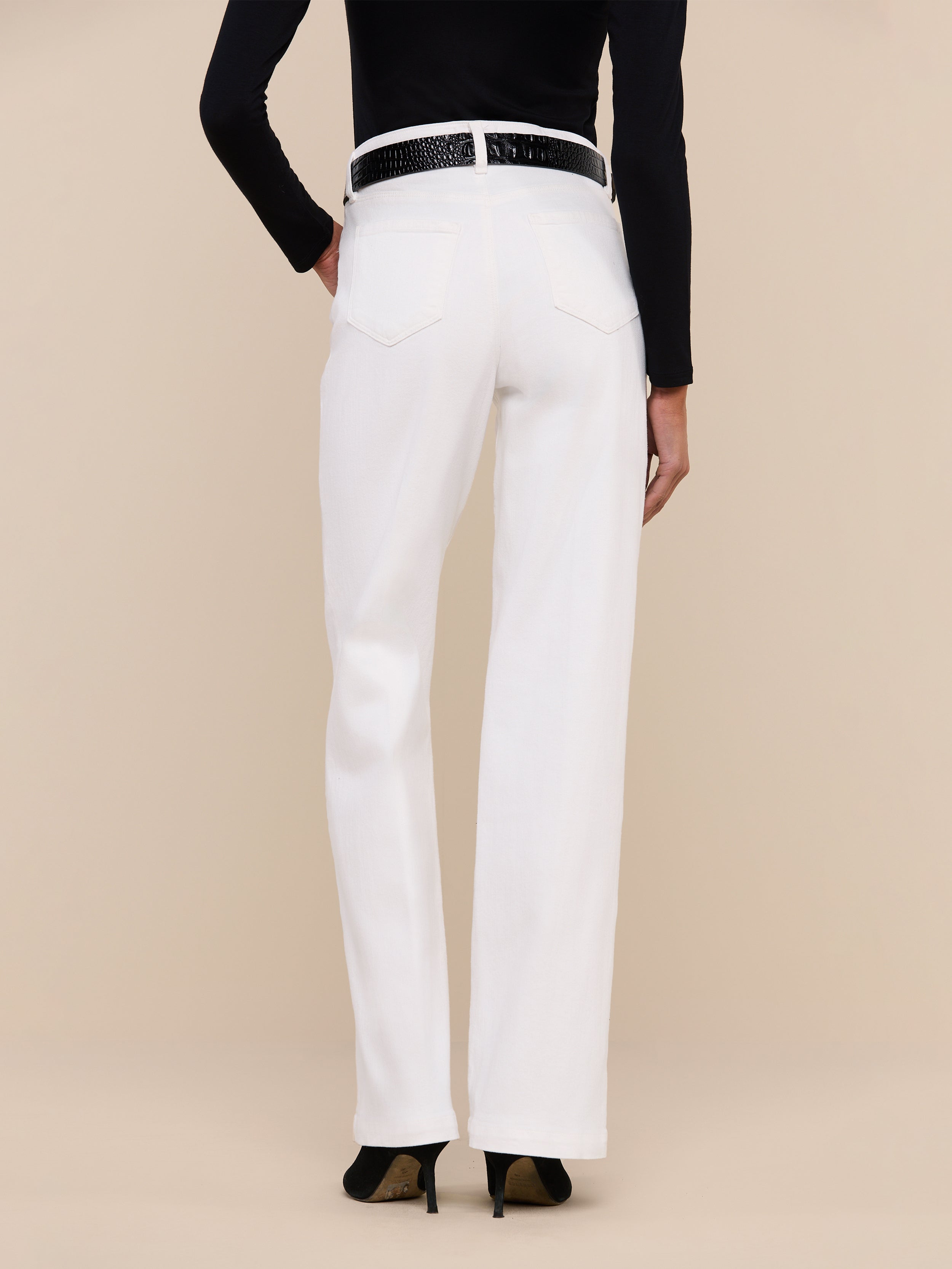 A person stands facing away from the camera, wearing the Scottie Wide-Leg Jean in white, featuring a high-rise waistband. The jeans are styled with a black belt and black top against a plain white background.