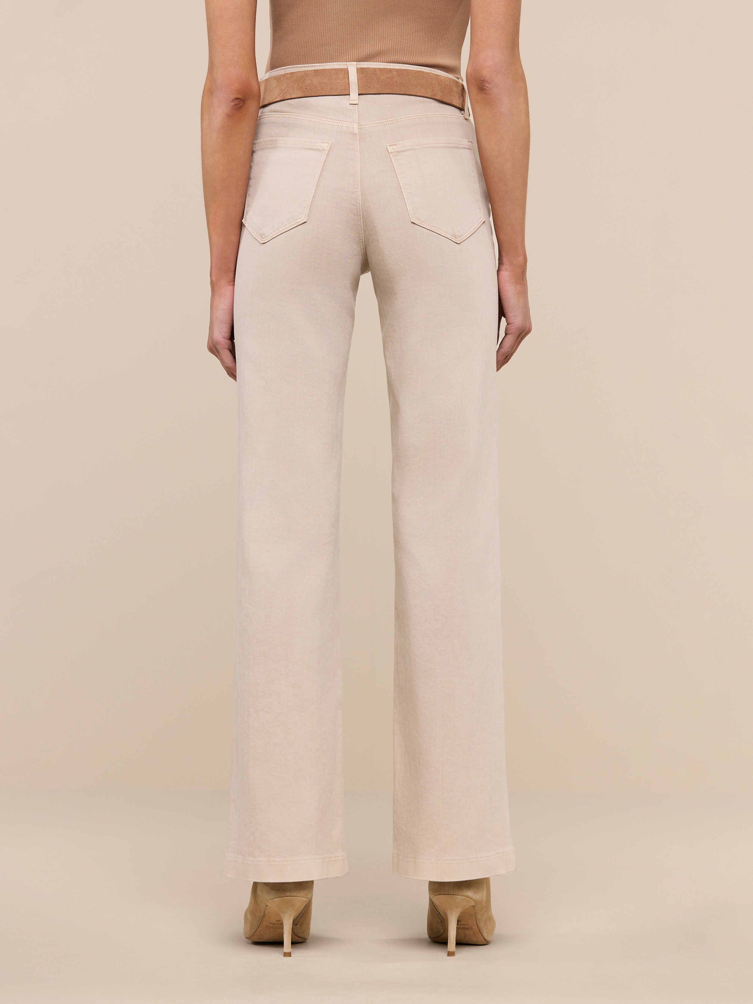 A person stands facing away, wearing the Scottie Wide-Leg Jean in a light beige wash with back pockets and tan high-heeled shoes against a plain white background.