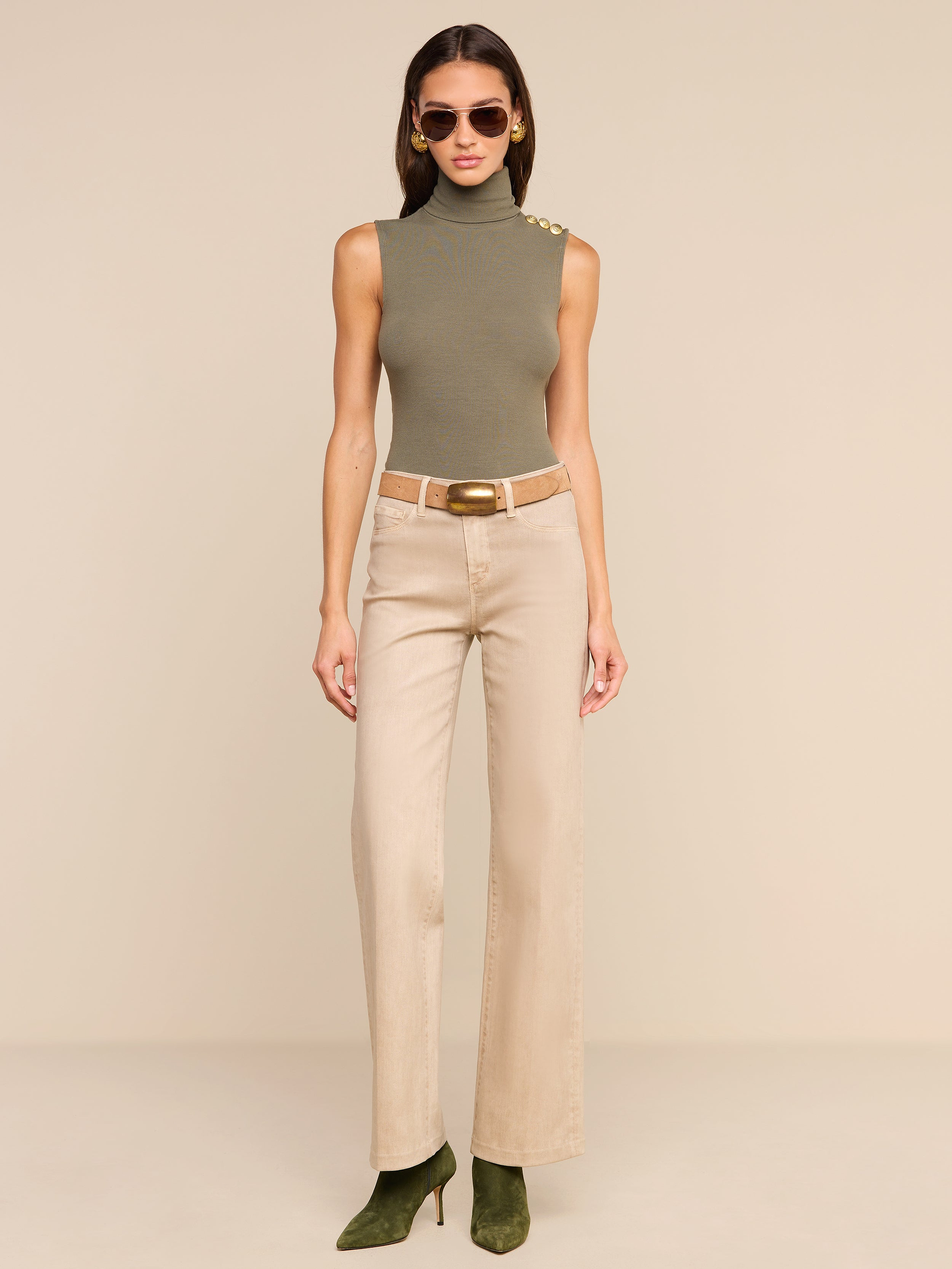 A woman in sunglasses stands against a plain white background, wearing a sleeveless olive green turtleneck, green heels, and the Scottie Coated Wide-Leg Jean in beige with a matching belt.