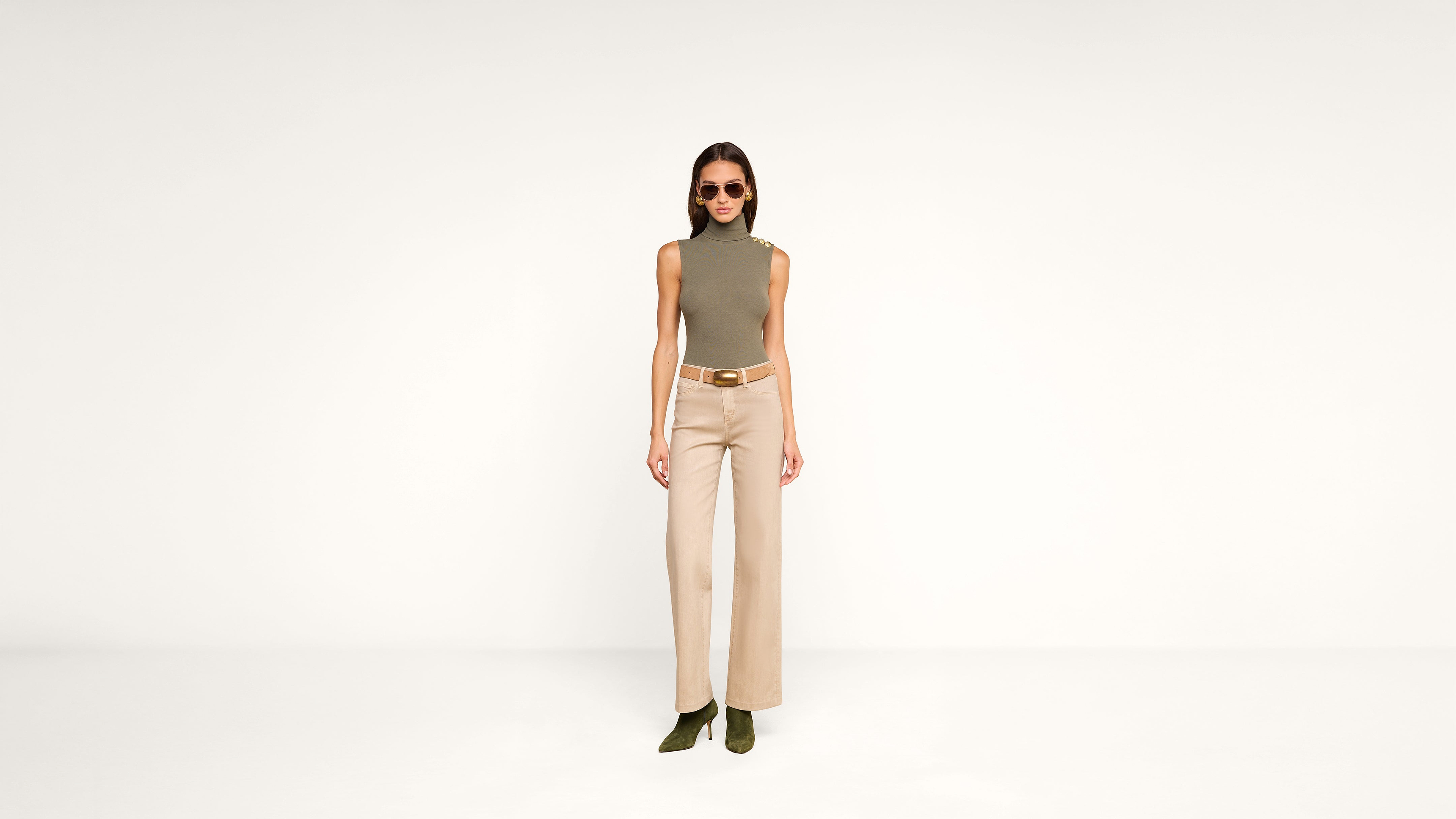 Woman wearing sunglasses, a sleeveless olive top, beige high-waisted wide-leg pants, a brown belt, and green heeled boots stands against a plain white background.