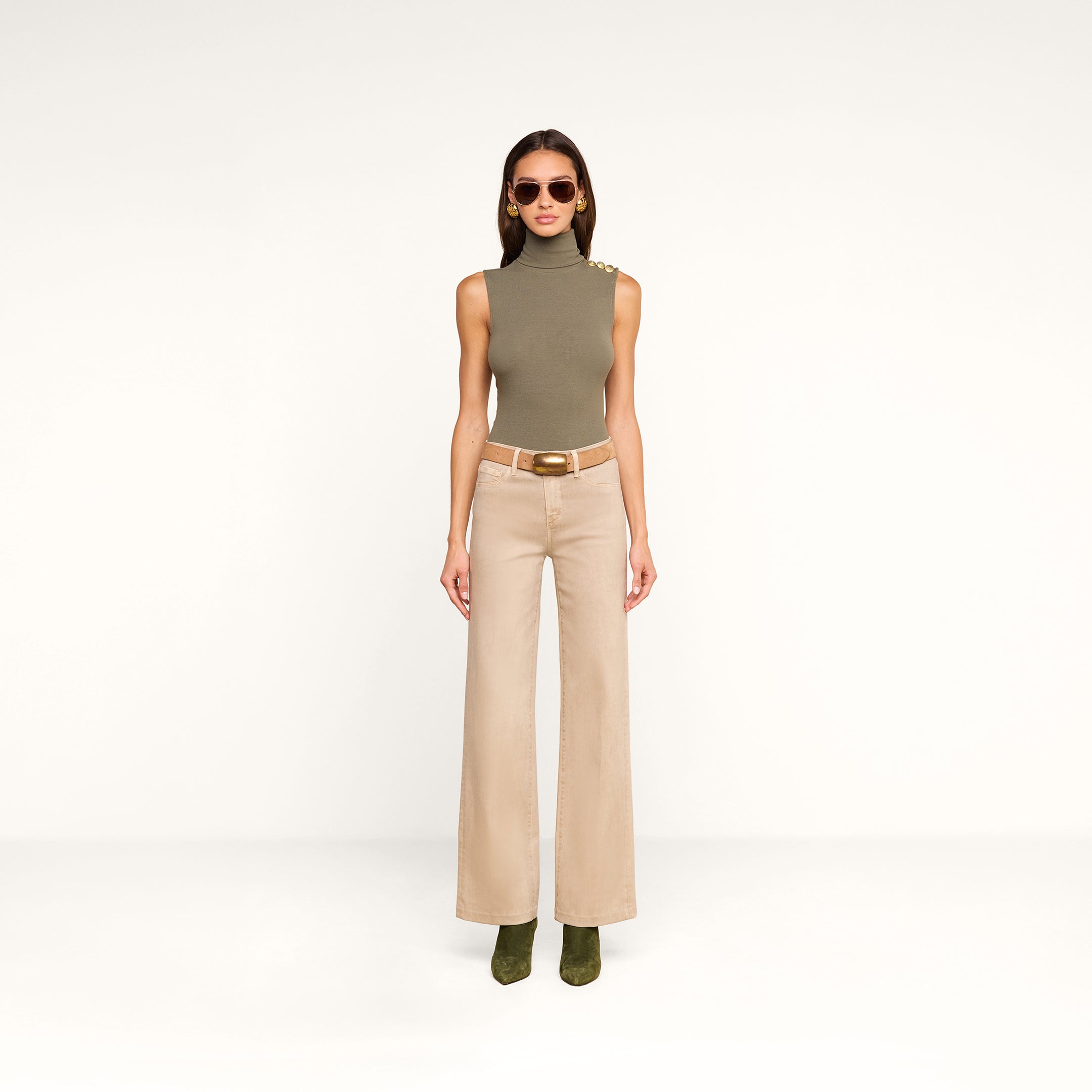 A woman in a sleeveless, olive-green turtleneck top, the Scottie Coated Wide-Leg Jean, green boots, and sunglasses stands against a plain white background.
