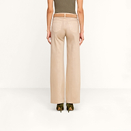 A person stands facing away, wearing the Scottie Coated Wide-Leg Jean in beige, paired with a tan belt, an olive green sleeveless top, and green heeled shoes against a plain white background.