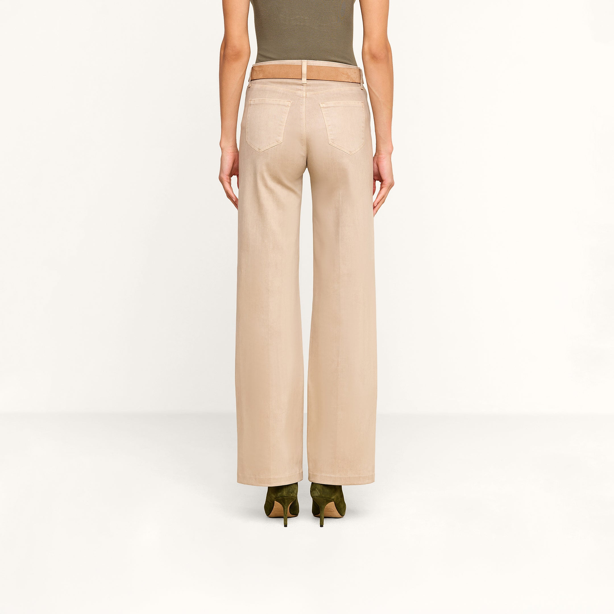 A person stands facing away, wearing the Scottie Coated Wide-Leg Jean in beige, paired with a tan belt, an olive green sleeveless top, and green heeled shoes against a plain white background.