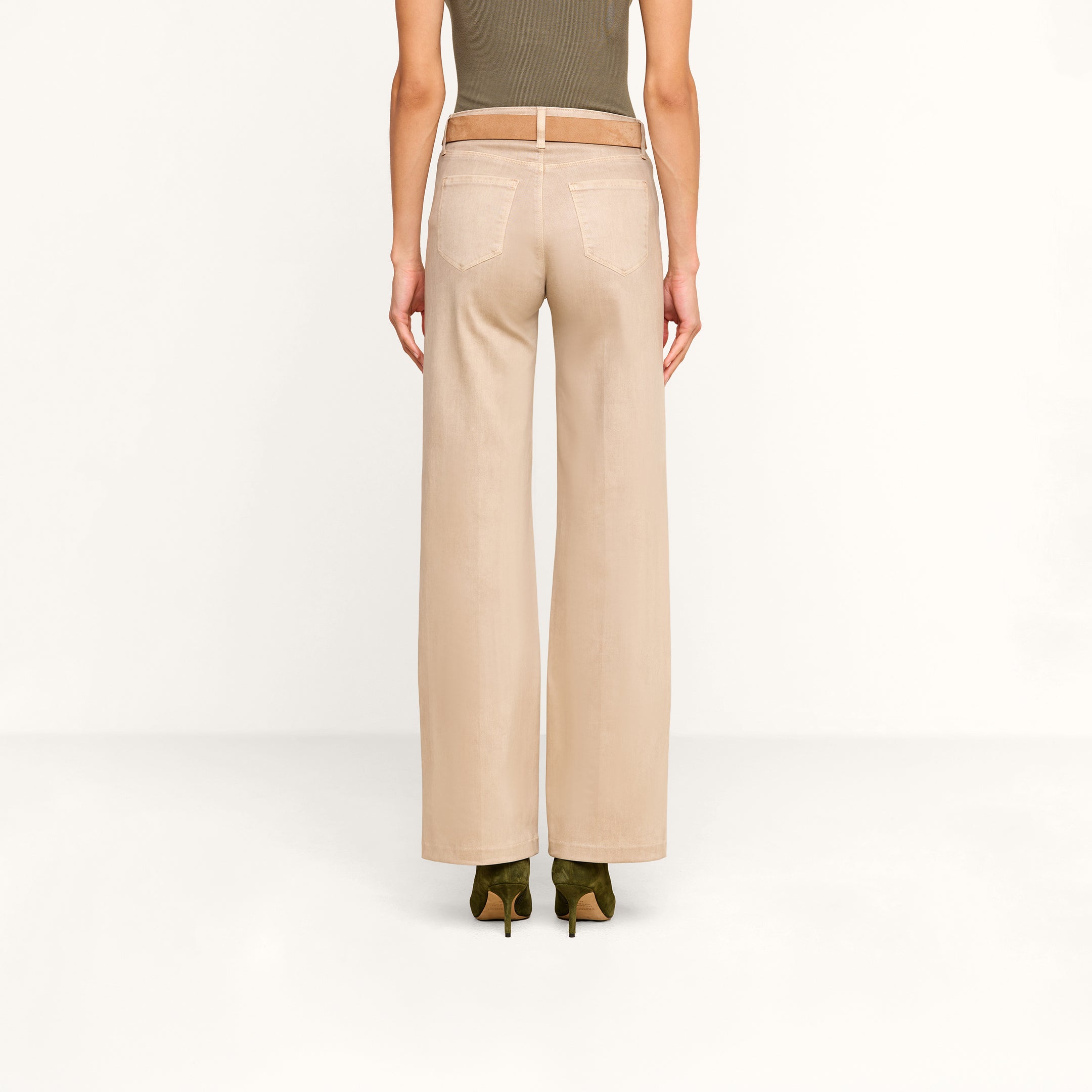 A person stands facing away, wearing the Scottie Coated Wide-Leg Jean in beige, paired with a tan belt, an olive green sleeveless top, and green heeled shoes against a plain white background.