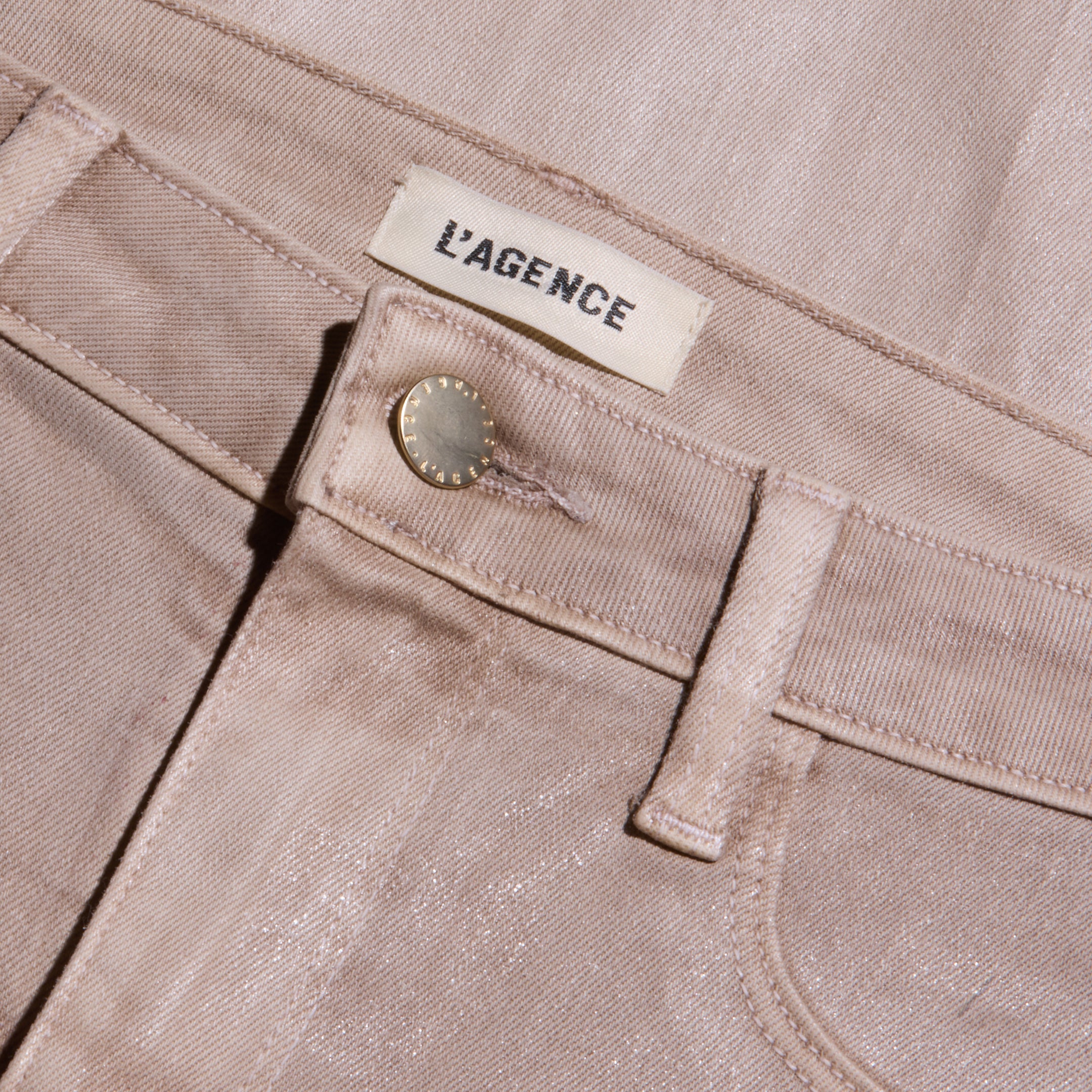 Close-up of the Scottie Coated Wide-Leg Jean in light pink, featuring a metallic button and a white L’AGENCE label sewn onto the waistband. The coated denim fabric gives off a subtle sheen, with visible stitching details.