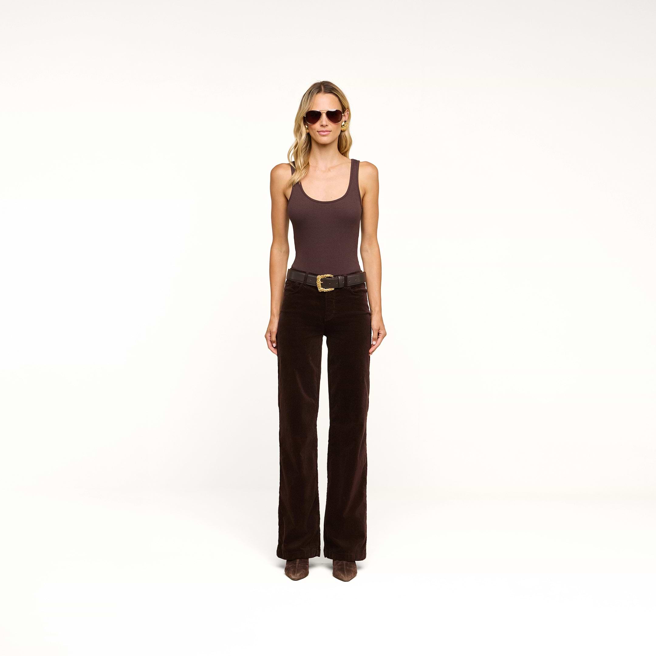 A woman wearing sunglasses, a brown sleeveless top, the Scottie Velvet Wide-Leg Jean—a pair of dark brown high-rise velvet pants with a contoured waistband—a belt, and brown boots stands against a plain white background.