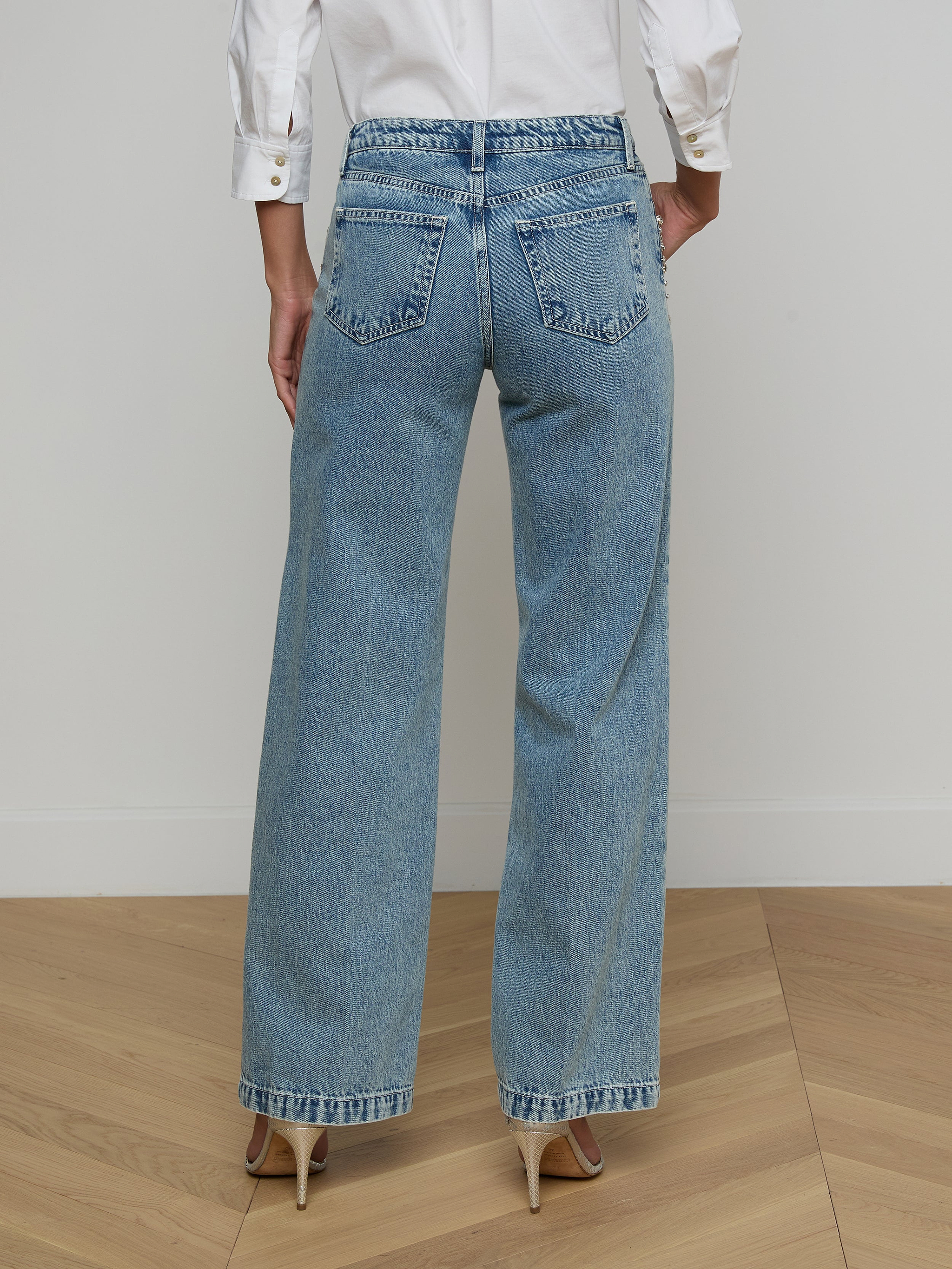 A person wearing a white long-sleeve shirt, beige high heels, and the Scottie Embellished Wide-Leg Jean in blue all-cotton denim is standing with their back to the camera against a plain light background.