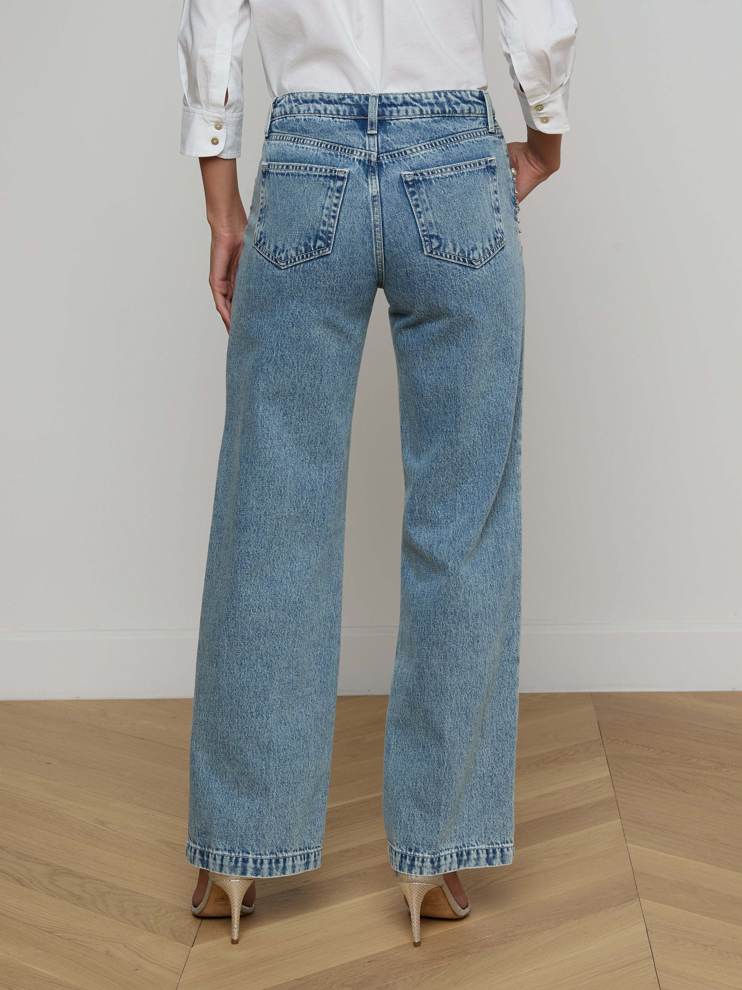 A person wearing a white long-sleeve shirt, beige high heels, and the Scottie Embellished Wide-Leg Jean in blue all-cotton denim is standing with their back to the camera against a plain light background.