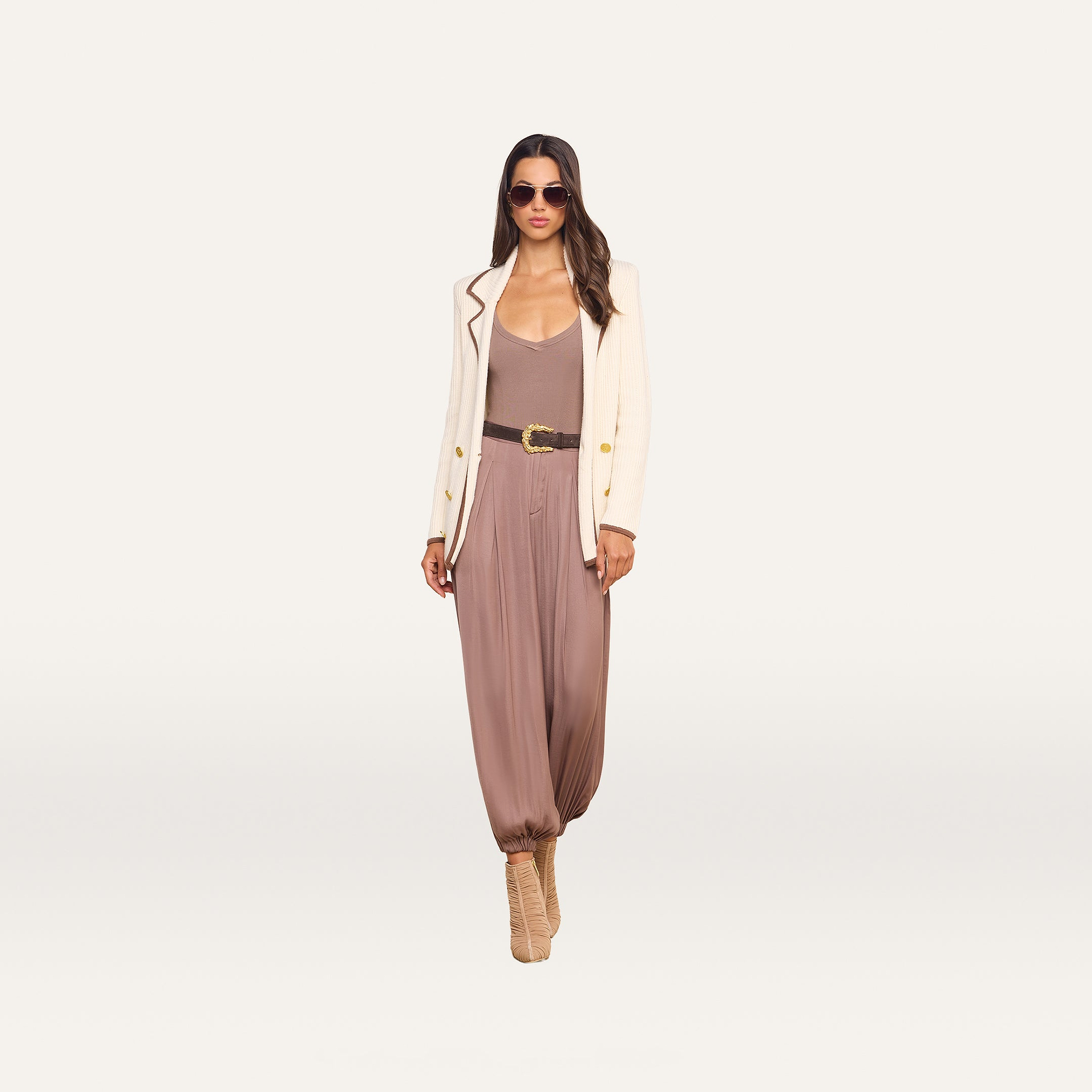 A woman stands against a plain white background, wearing sunglasses, a cream blazer, the Palmer Satin Pant styled with a belt, and beige high-heeled shoes.