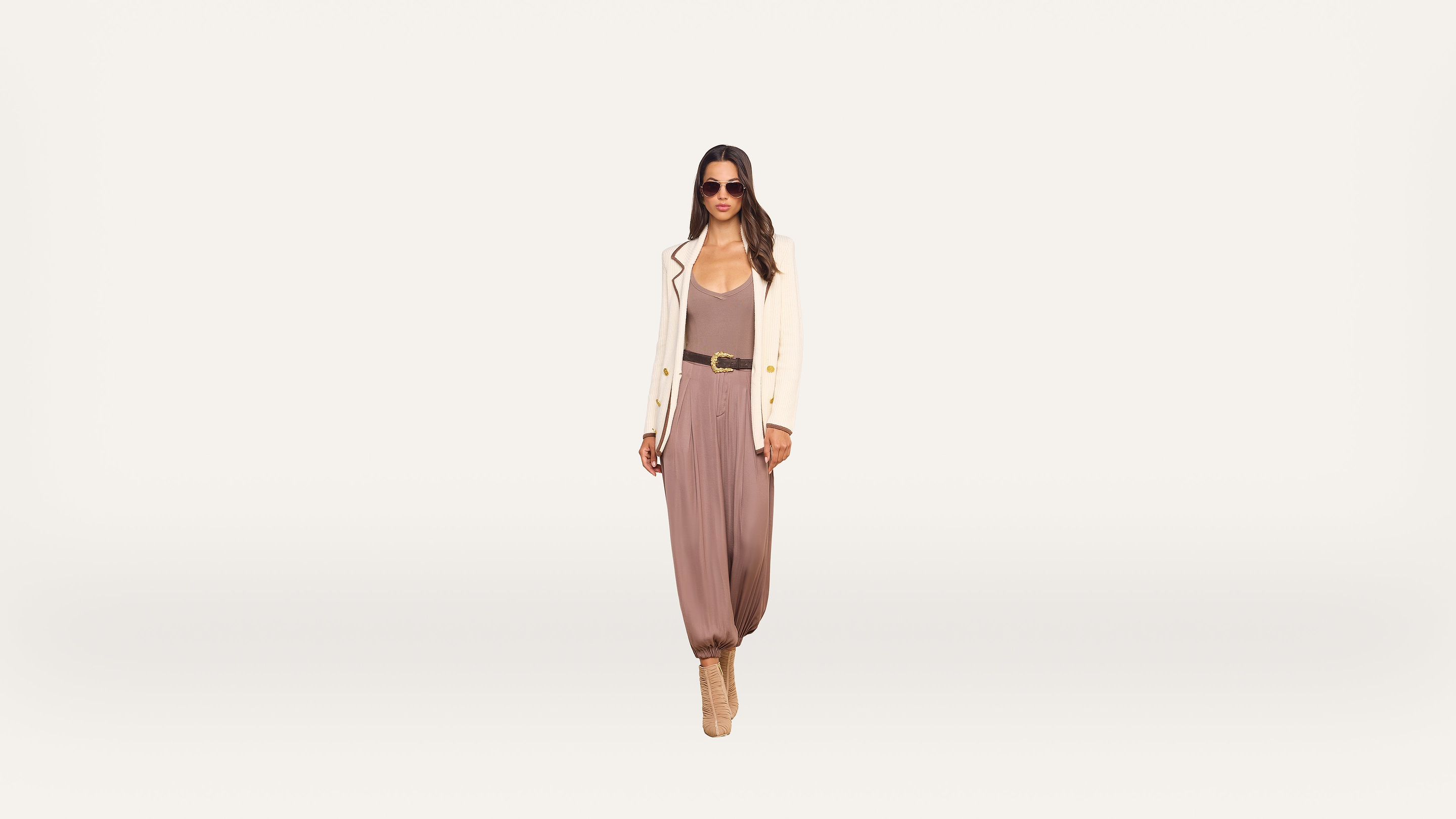 A woman in sunglasses wears a cream blazer, taupe jumpsuit, black belt, and high-heeled boots, walking confidently against a plain white background.