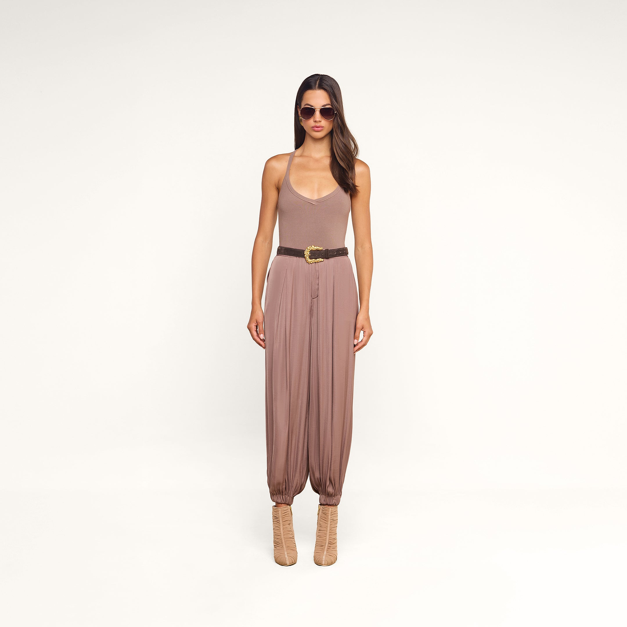 A woman stands against a white background wearing sunglasses, a taupe tank top, the Palmer Satin Pant—a pair of high rise loose brown balloon trousers—styled with a black belt and beige ankle boots.