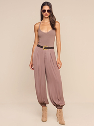A woman wearing sunglasses, a taupe tank top, the high-rise loose Palmer Satin Pant in mauve with a belt, and tan ankle boots stands against a plain white background.