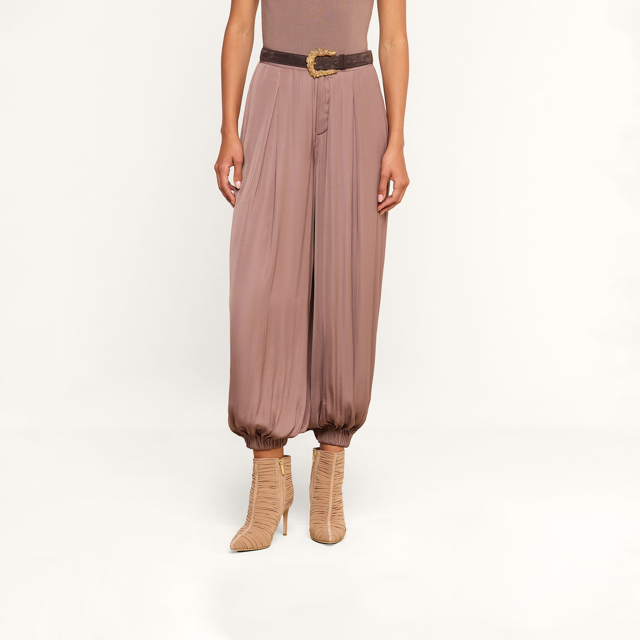 A person wearing the Palmer Satin Pant—a loose, mauve high-rise balloon trouser styled with a dark belt—paired with a taupe sleeveless top and beige heeled boots, stands against a white background.