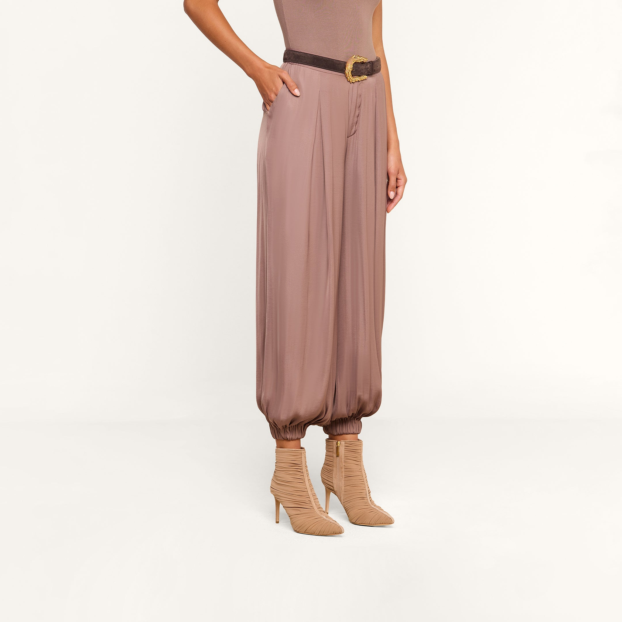 A person wearing the Palmer Satin Pant in taupe, styled with a brown belt and beige heeled ankle boots, stands against a plain white background.