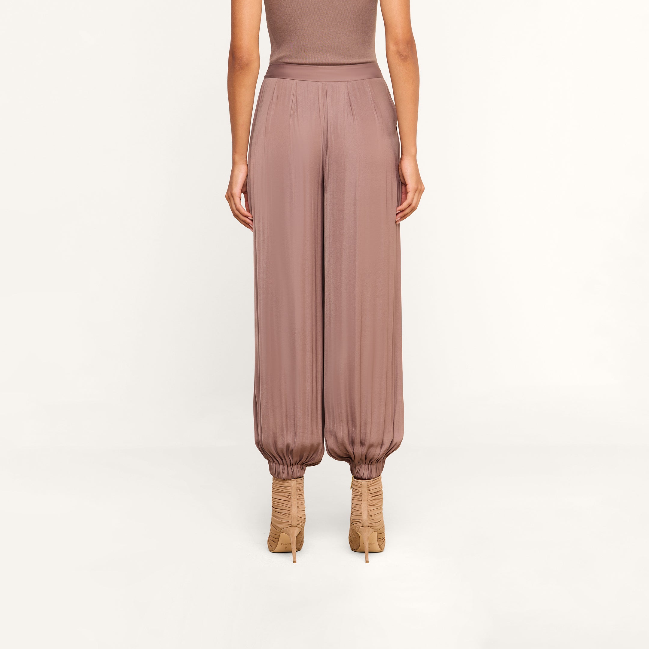A woman stands facing away, wearing the Palmer Satin Pant in mauve—a high-rise balloon trouser that tapers at the ankles—paired with beige high-heeled shoes, against a plain light background.