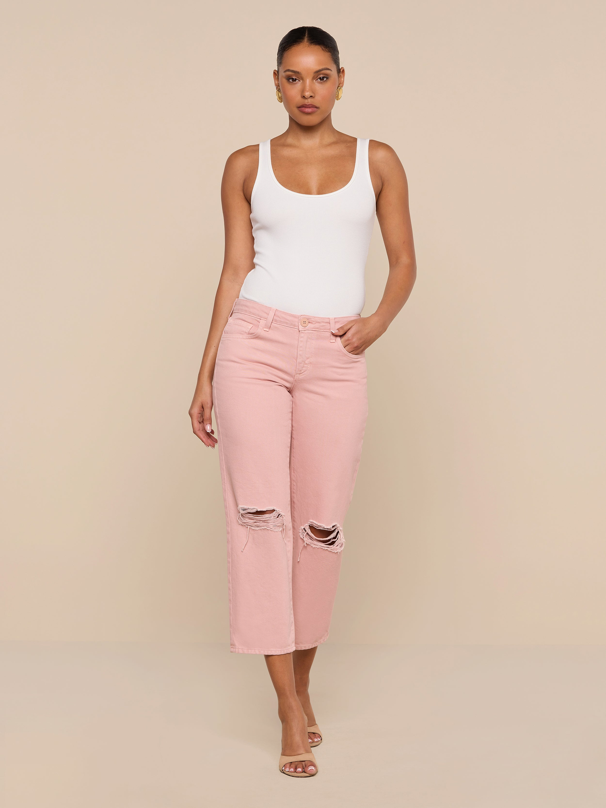 A woman stands against a plain white background wearing a white tank top, the Blaine Cropped Stovepipe Jean in light pink with a straight-leg fit and low rise, and heeled sandals, with one hand in her pocket.
