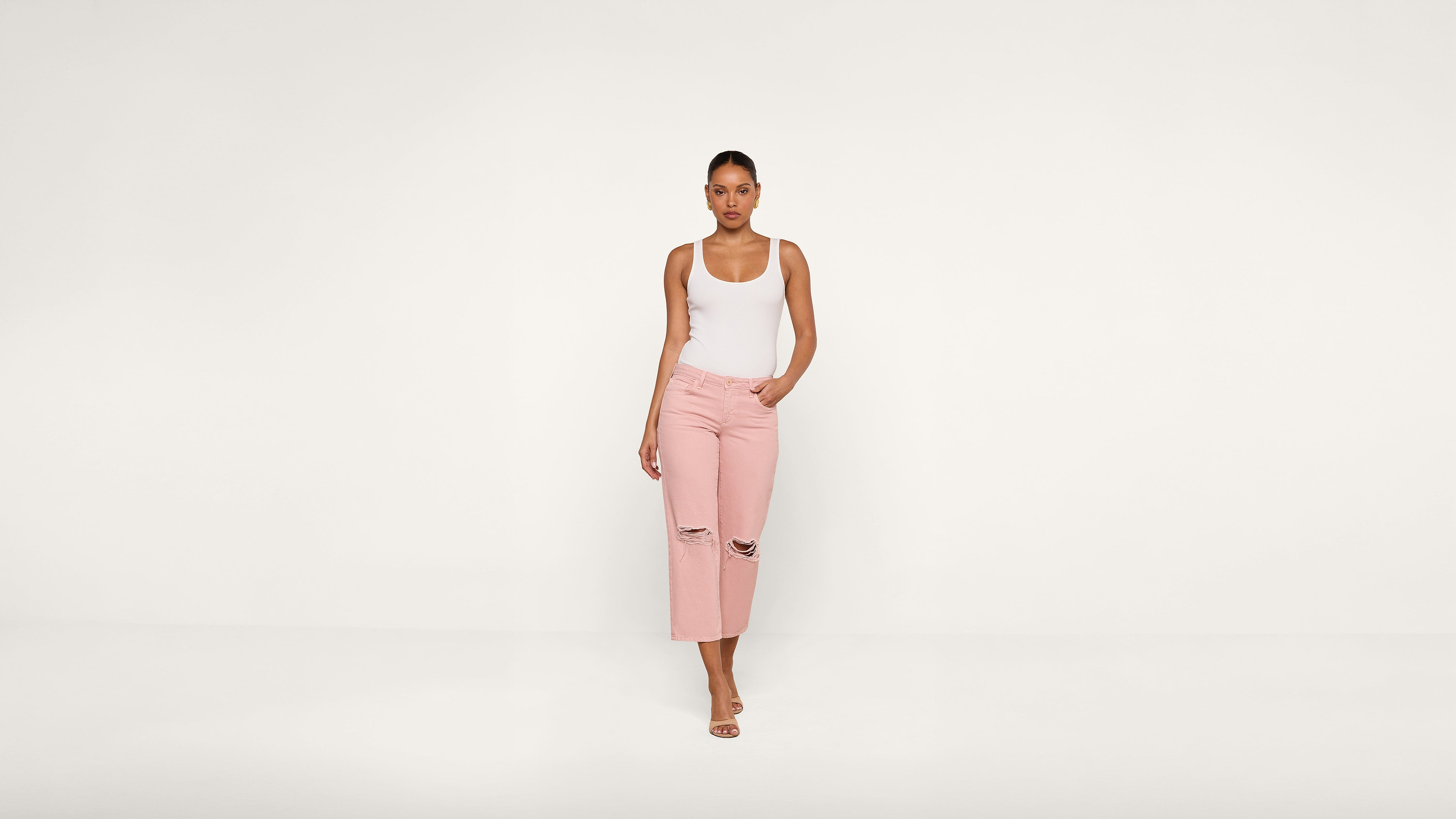 A woman stands against a plain white background, wearing a white tank top, light pink ripped jeans, and sandals, with one hand in her pocket.