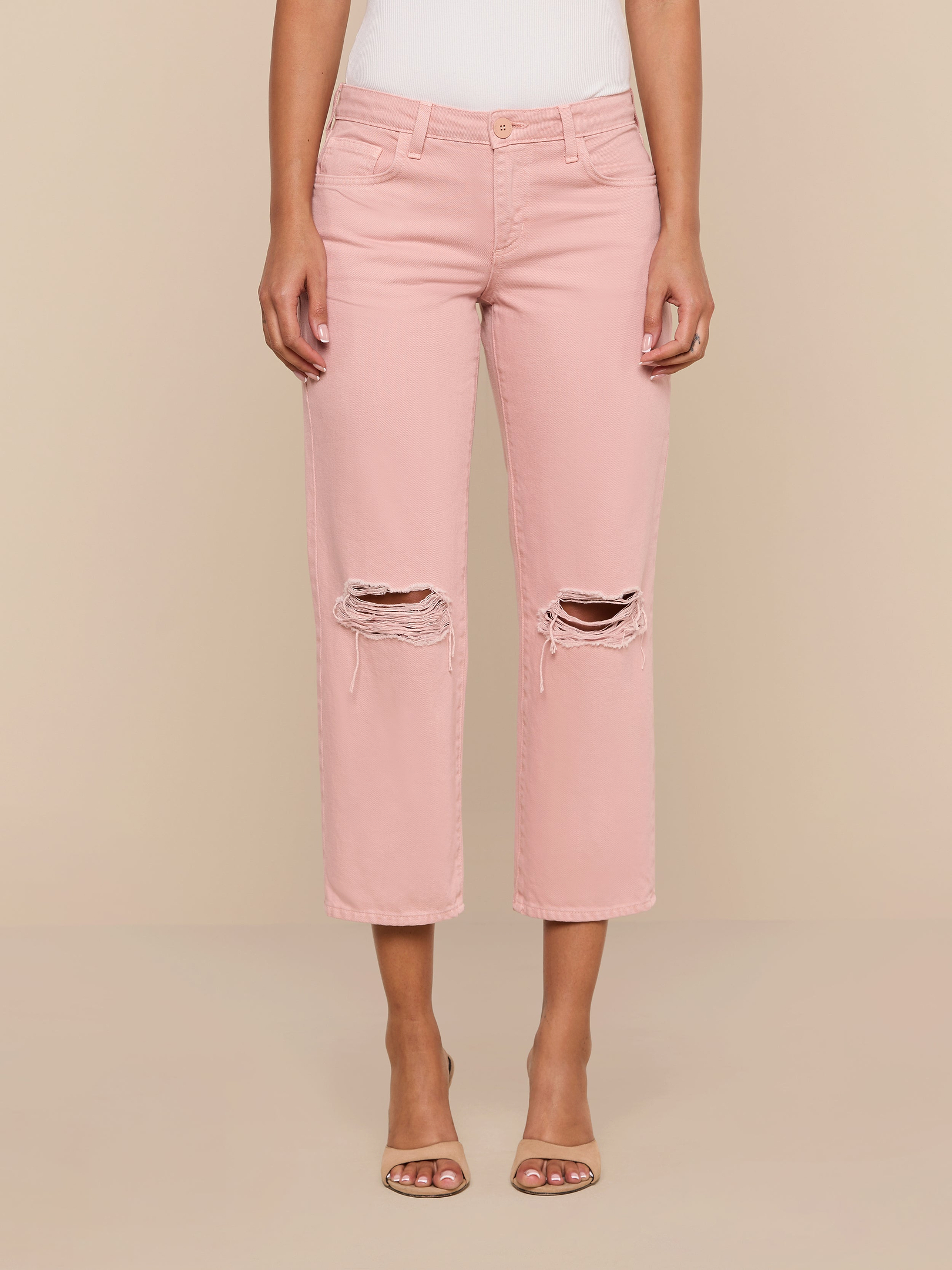 A person wearing the Blaine Cropped Stovepipe Jean in light pink with a ripped, low-rise, straight-leg fit and open-toed heels stands against a plain white background.