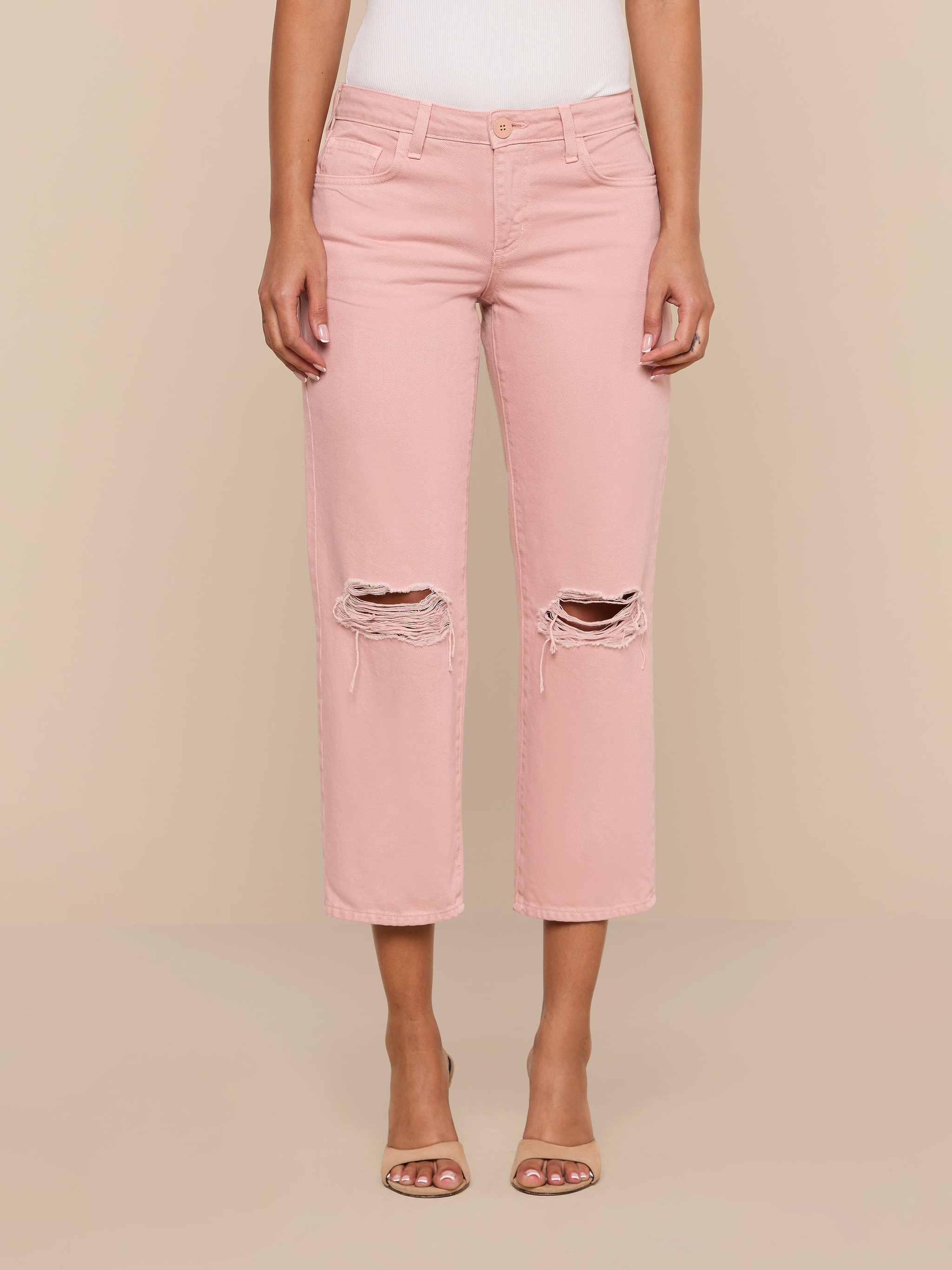 A person wearing the Blaine Cropped Stovepipe Jean in light pink with a ripped, low-rise, straight-leg fit and open-toed heels stands against a plain white background.