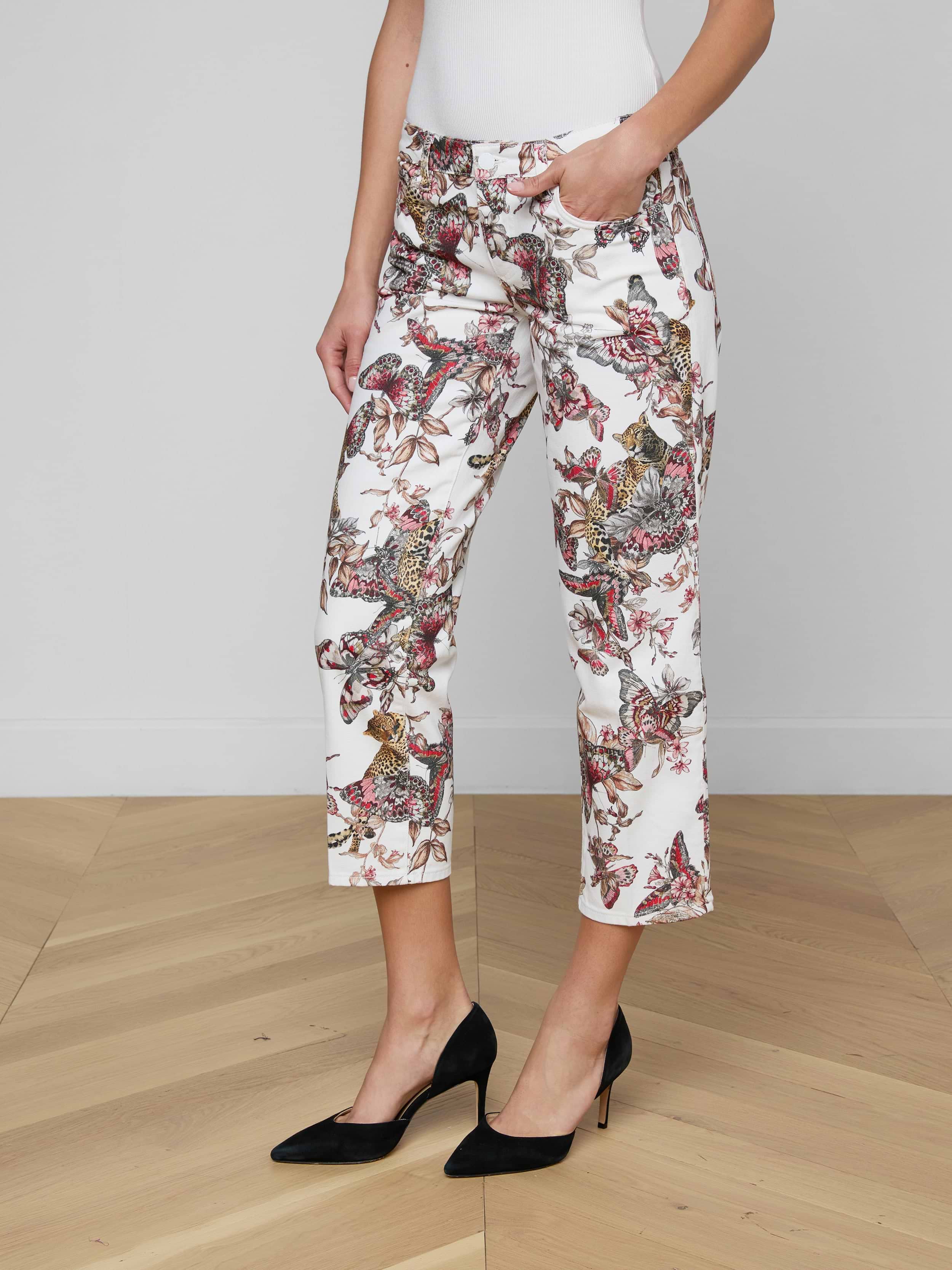 A woman stands against a plain background wearing the Blaine Cropped Stovepipe Jean—white, straight-leg, ankle-length jeans featuring a colorful butterfly print—paired with a white top and black high heels, with one hand in her pocket.