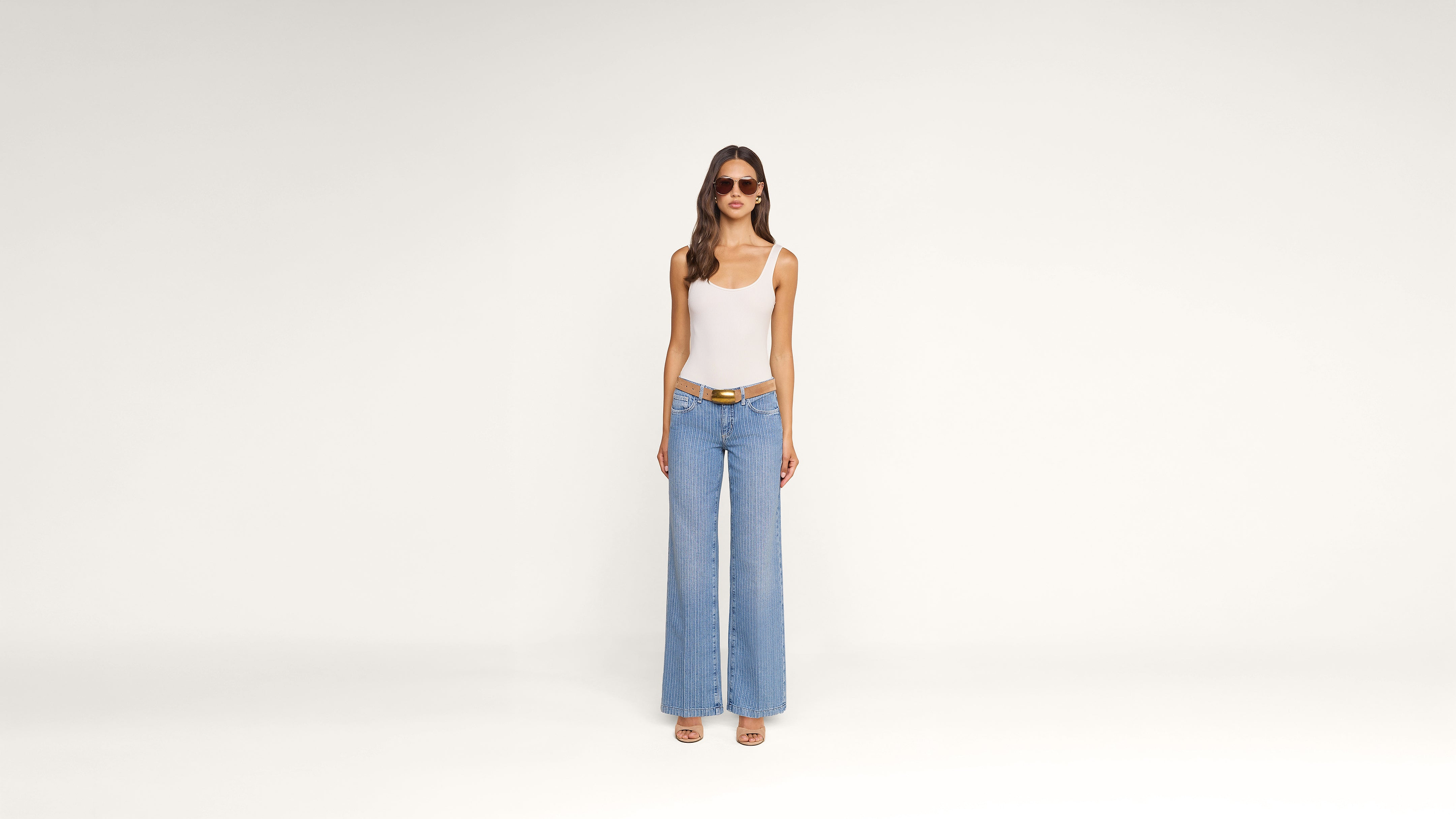 A woman wearing sunglasses, a white tank top, blue wide-leg jeans, a brown belt, and sandals stands against a plain white background.