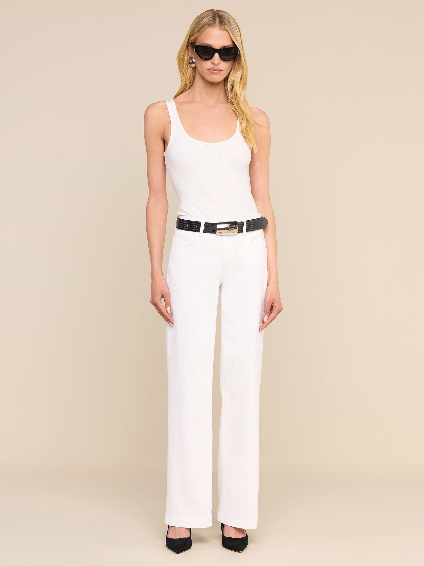 Une femme portant des lunettes de soleil noires, un débardeur blanc et un jean chic « Joanne Low-Rise Wide-Leg » agrémenté dune ceinture noire et de talons noirs se tient debout devant un fond beige uni.